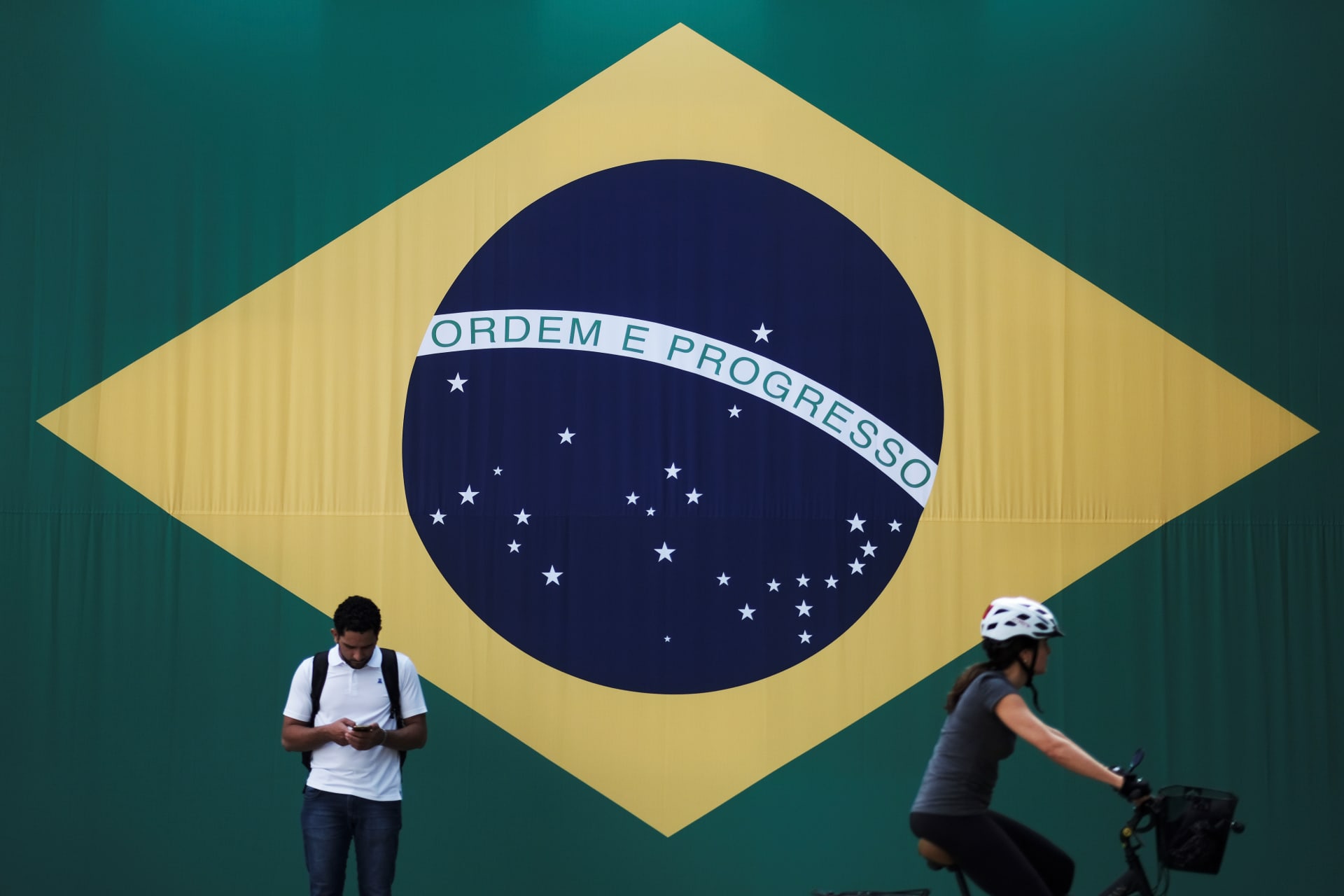 <p>A man checks his mobile phone as a woman riding a bike passes next to a big Brazilian flag in Sao Paulo, Brazil June 28, 2018. </p>