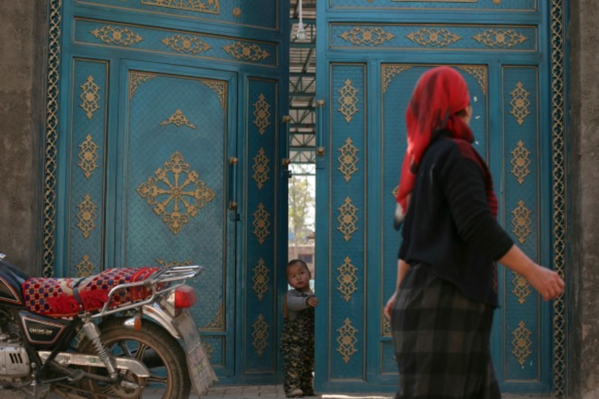 <p>A child looks out from a door as a Uighur woman walks by in a residential area in Turpan, Xinjiang Uighur Autonomous Region on October 31, 2013 (Michael Martina/Courtesy Reuters).</p>
