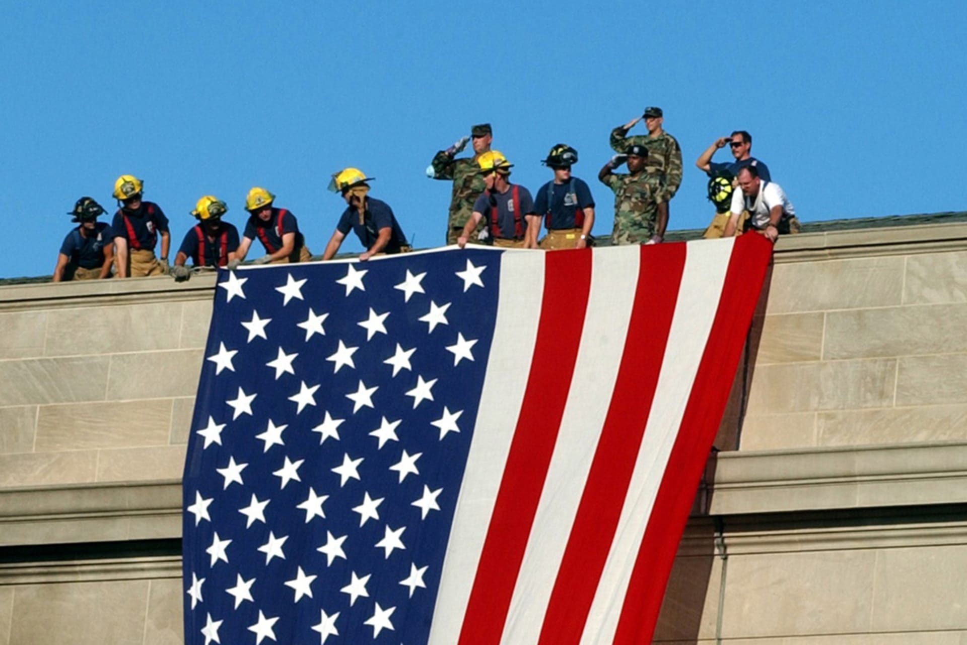 <p>Military service members salute as fire and rescue workers unfurl a huge American flag over the side of the Pentagon on Sept. 12, 2001.</p>