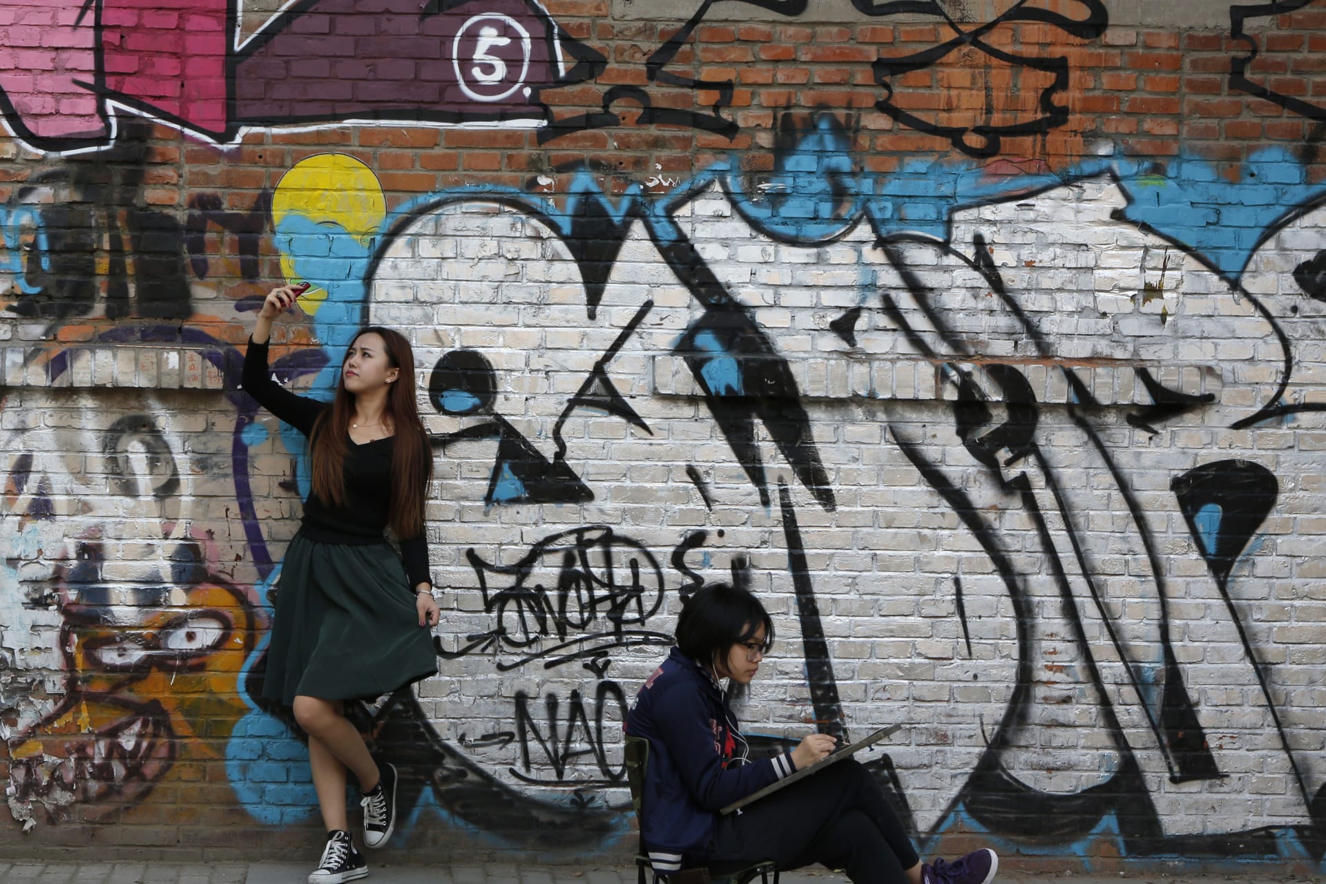 <p>A woman poses for her self-portrait with her mobile phone while another works on a painting in front of a graffiti during the 2013 Beijing 798 Art Festival at the 798 Art Zone in Beijing October 8, 2013. The art zone, originally an unused factory, was tra</p>
