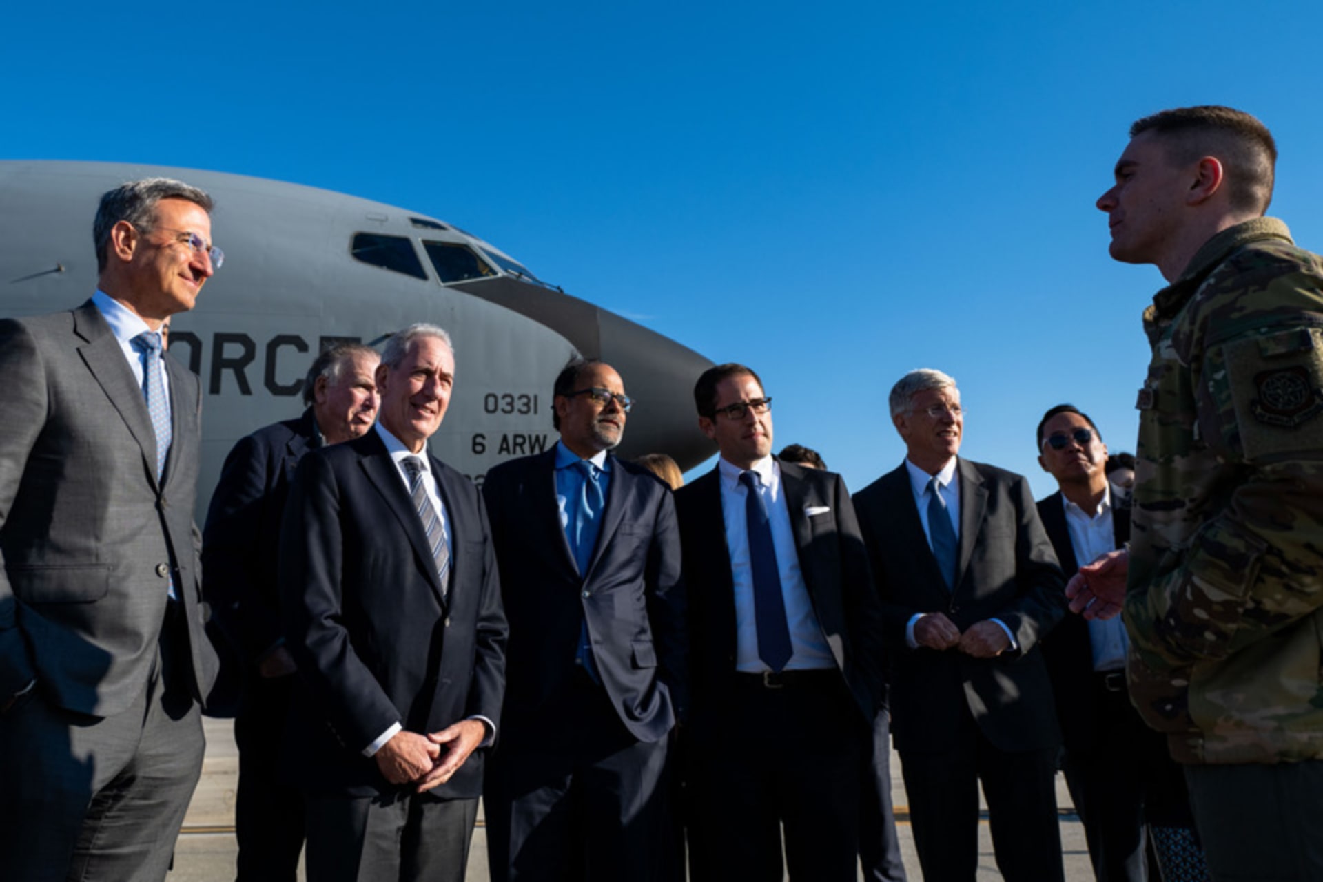 A group of men in suits stand in front of a grey military aircraft.