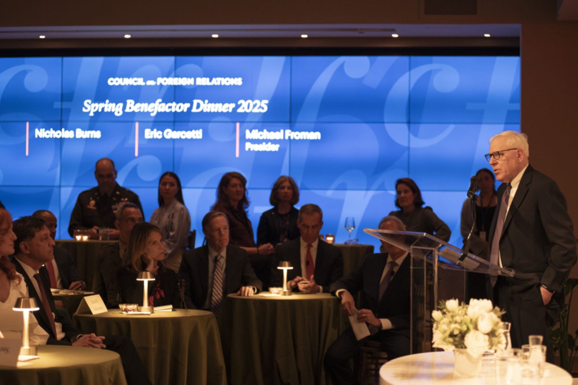 David Rubenstein stands in front of a sitting audience in a dark room with a blue screen behind them.