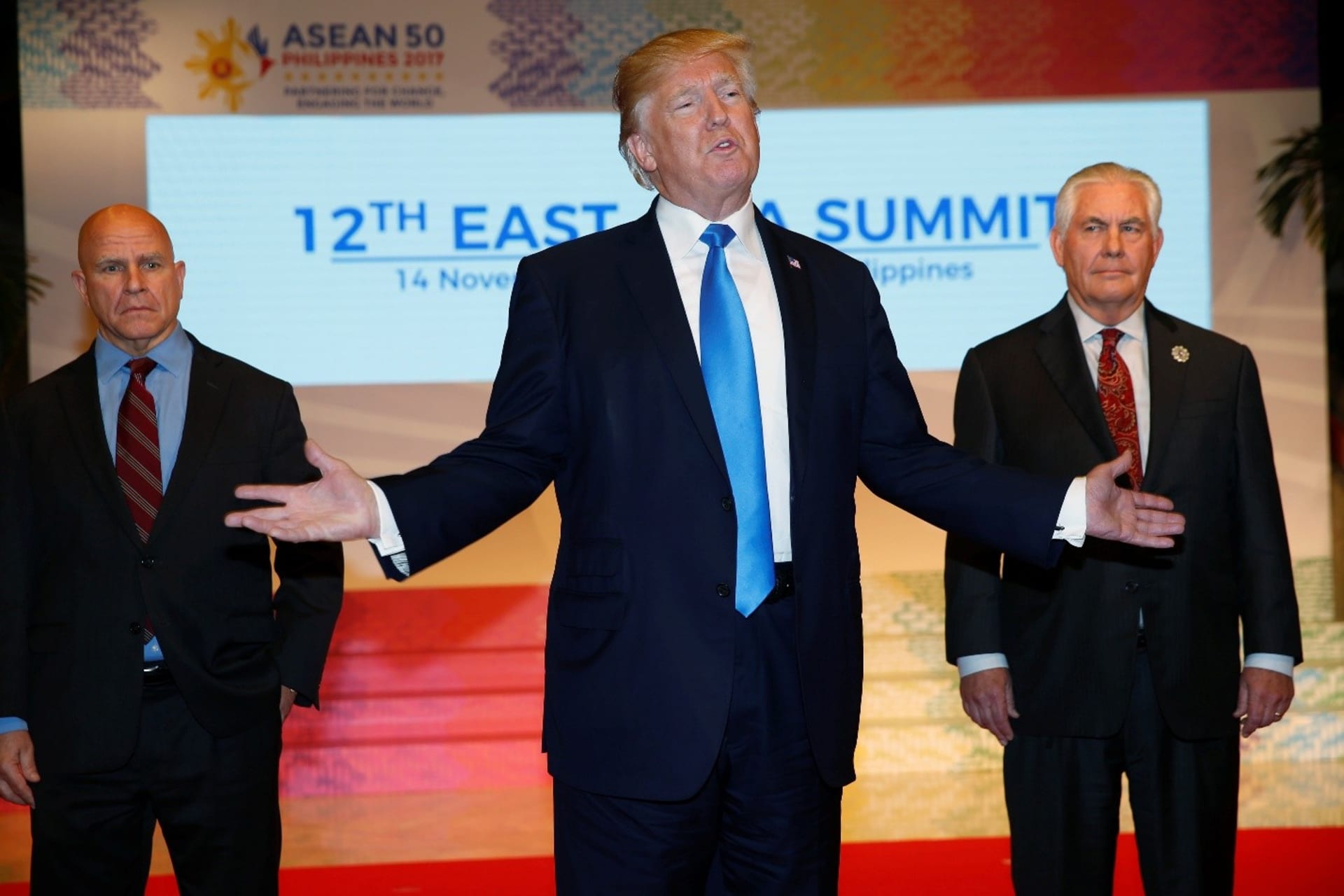 U.S. President Donald Trump stands between two aides in front of projected signage reading "12th East Asian Summit"