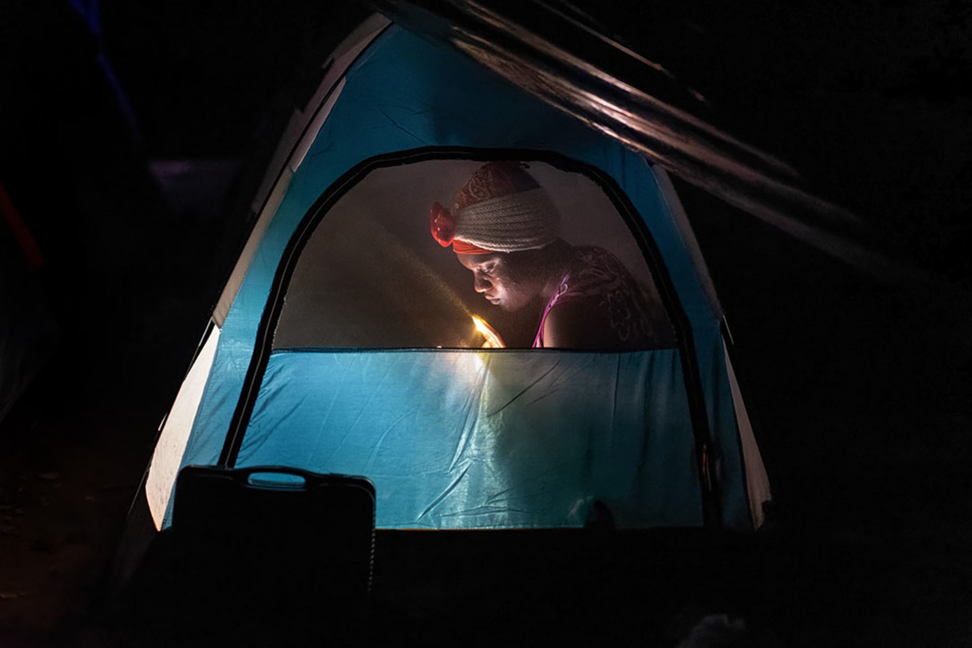 A woman immigrant from Haiti is seen through the screen of a tent reading by flashlight.