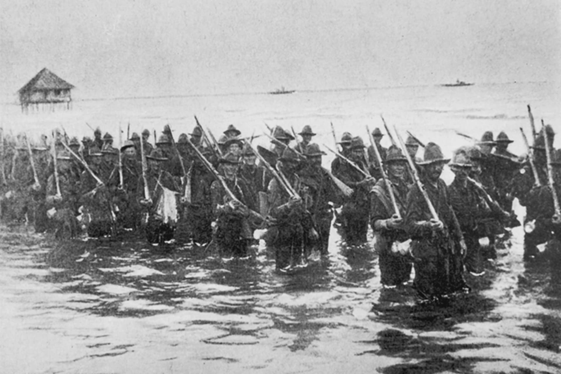 A black and white photo of American troops standing in the water on the shore of Manila holding rifles. 