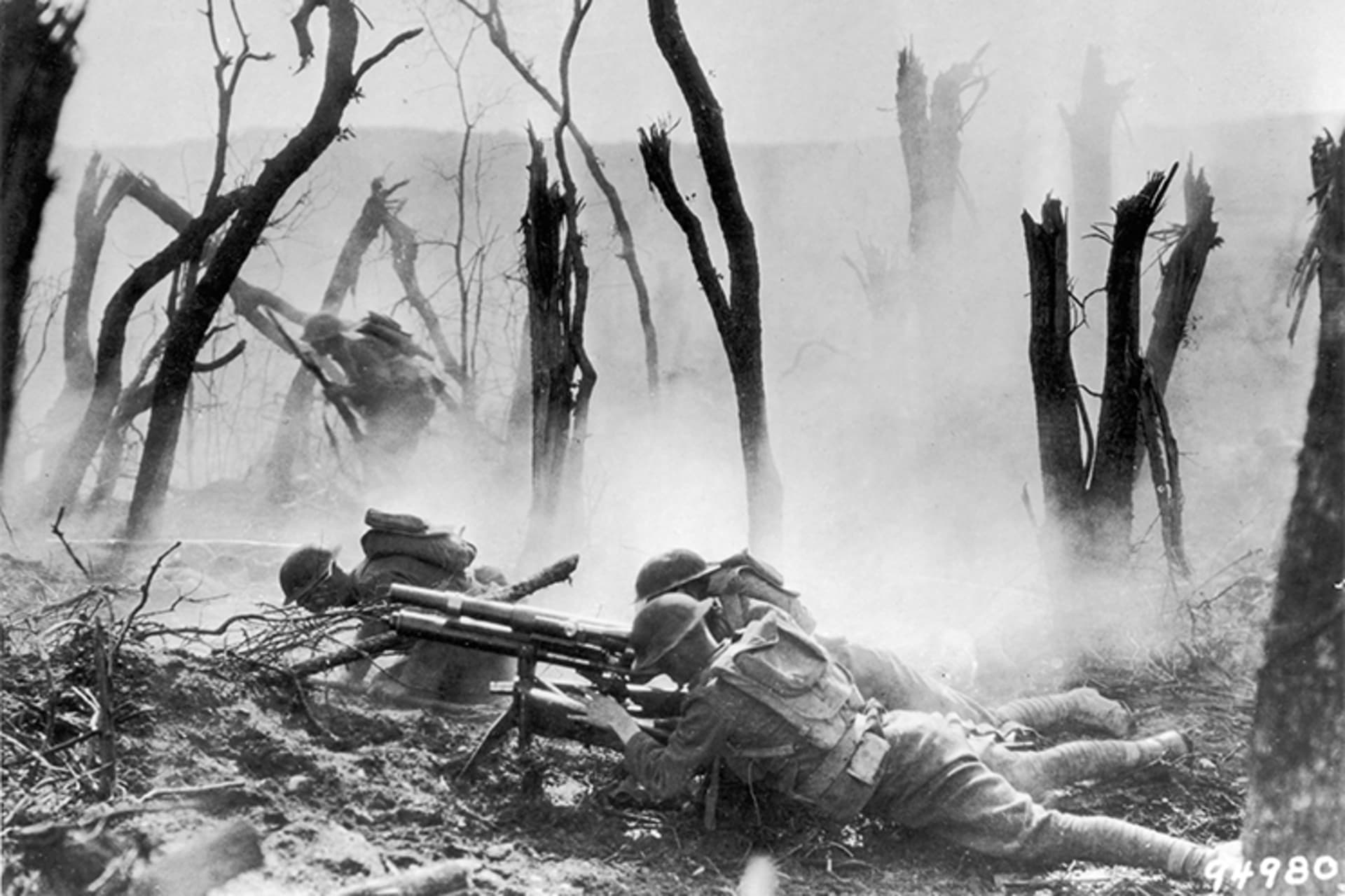 A black and white still from a documentary showing U.S. soldiers, surrounded by destroyed trees, laying down and firing a machine gun at German forces. 