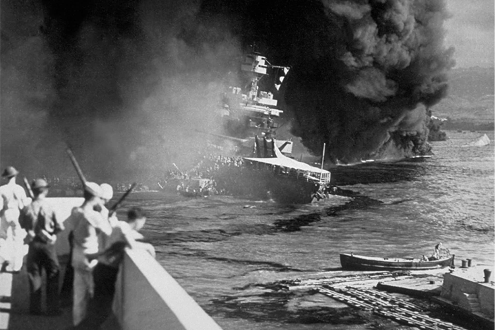 A black and white image of a U.S. ship on fire after the Pearl Harbor attack. Soldiers look on at the burning sight. 