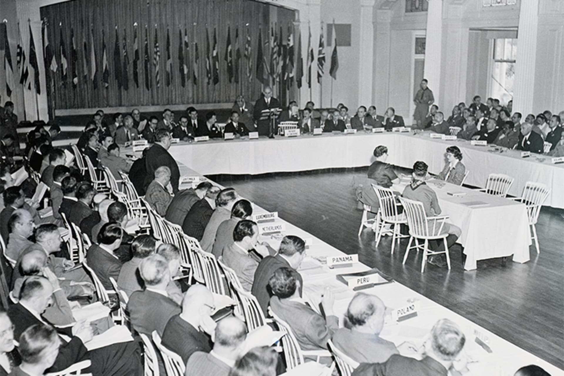 A black and white photo of members gathered at the Bretton Woods Conference. The background shows various United Nations country flags.