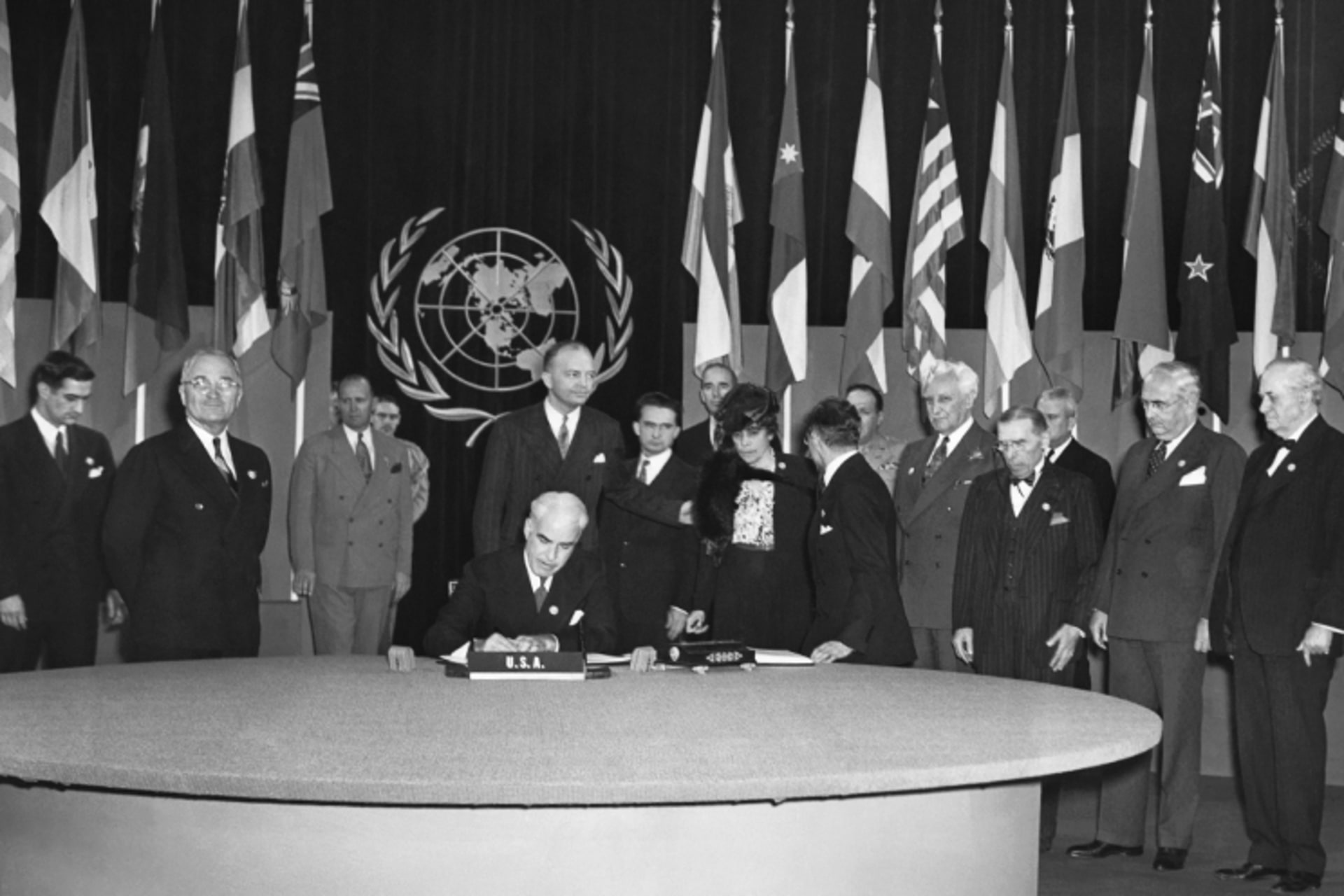A black and white photo of the signing of the UN Charter. The UN logo and member country flags are in the back.