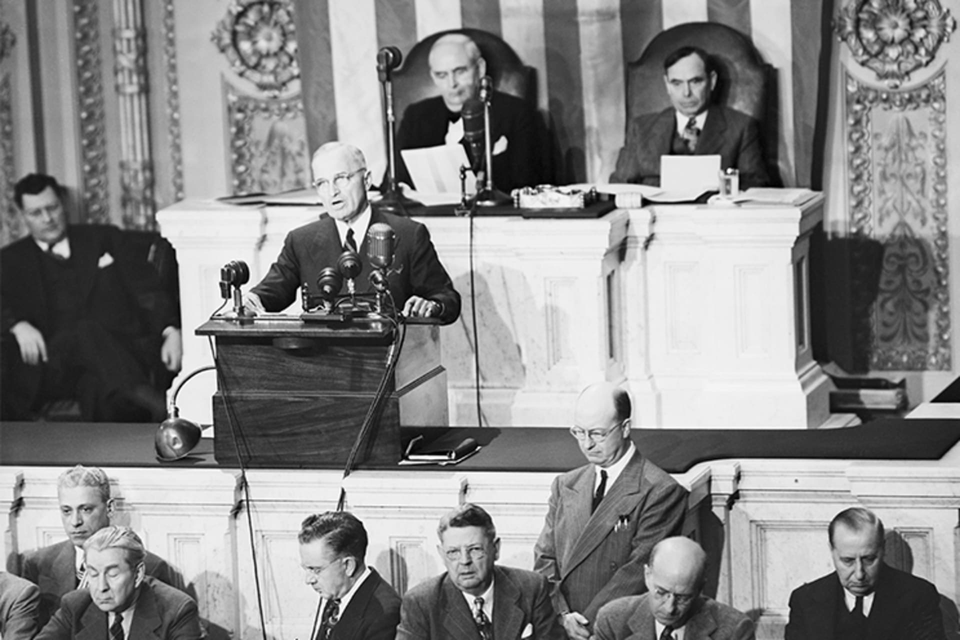 A black and white photo of President Truman addressing Congress from a podium with many mics. 