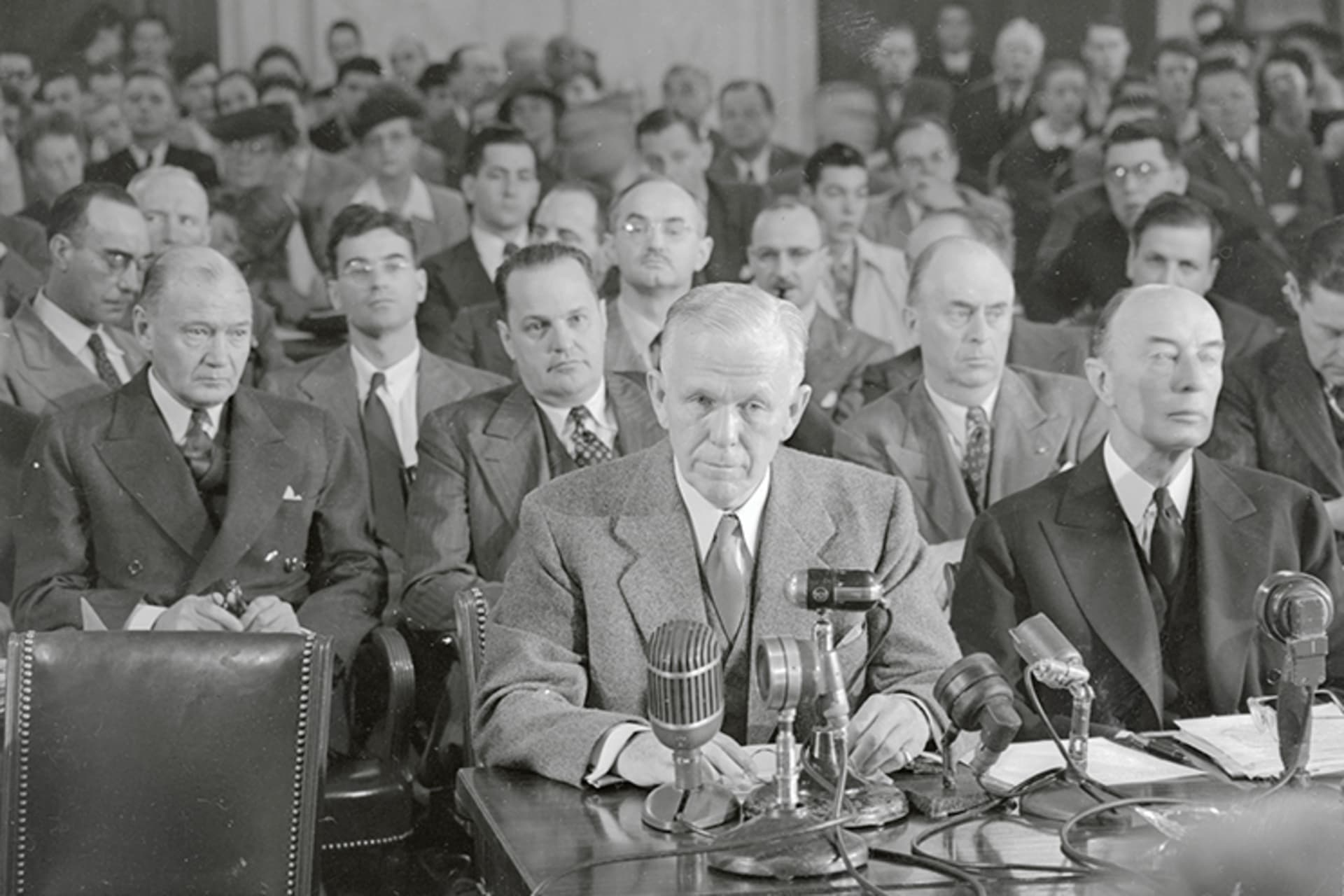 A black and white photo of Secretary of State George Marshall seated at a desk with many mics as he answers questions from the Senate.