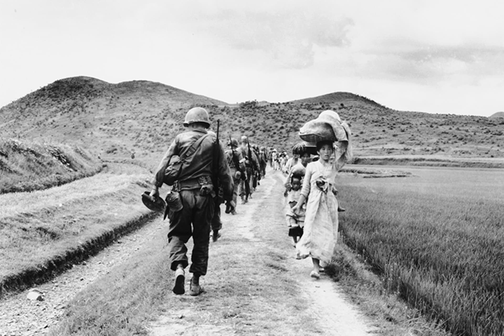 A black and white photo of a line of U.S. soldiers walking away from the camera on the left side and a line of Korean refugees walking towards the camera, carrying their belongings, on the right side.