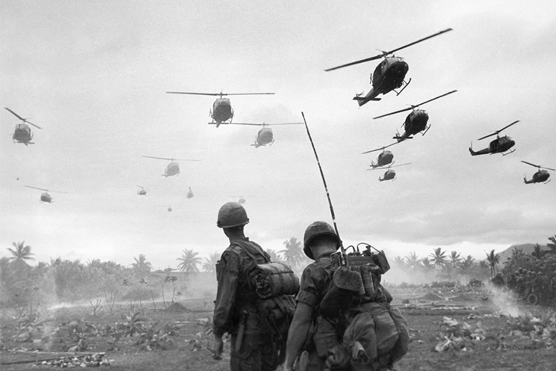 A black and white photo of many helicopters flying over two U.S. soldiers on the ground as they look up. 