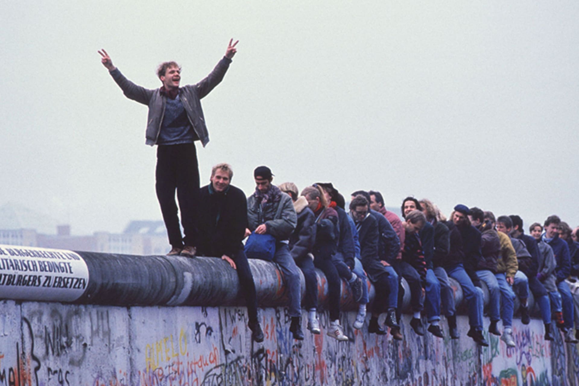 A photo of people atop the Berlin Wall celebrating the collapse of communism. The man at the front stands, while the rest of the group is seated, and raises his arms holding up two peace signs with his fingers. 