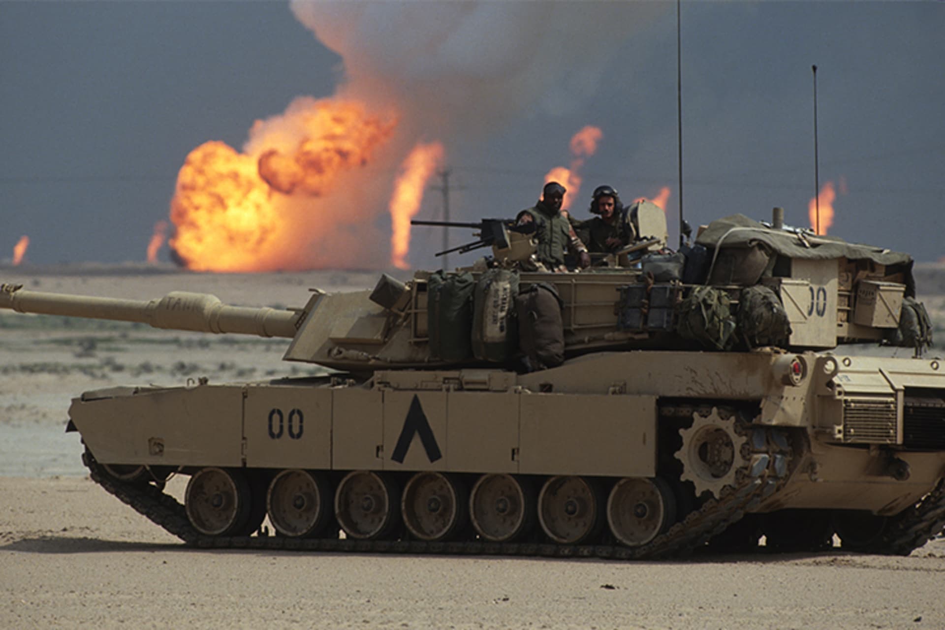 A photo of U.S. soldiers stop a tank as oil wells burn in the background and flames are seen.