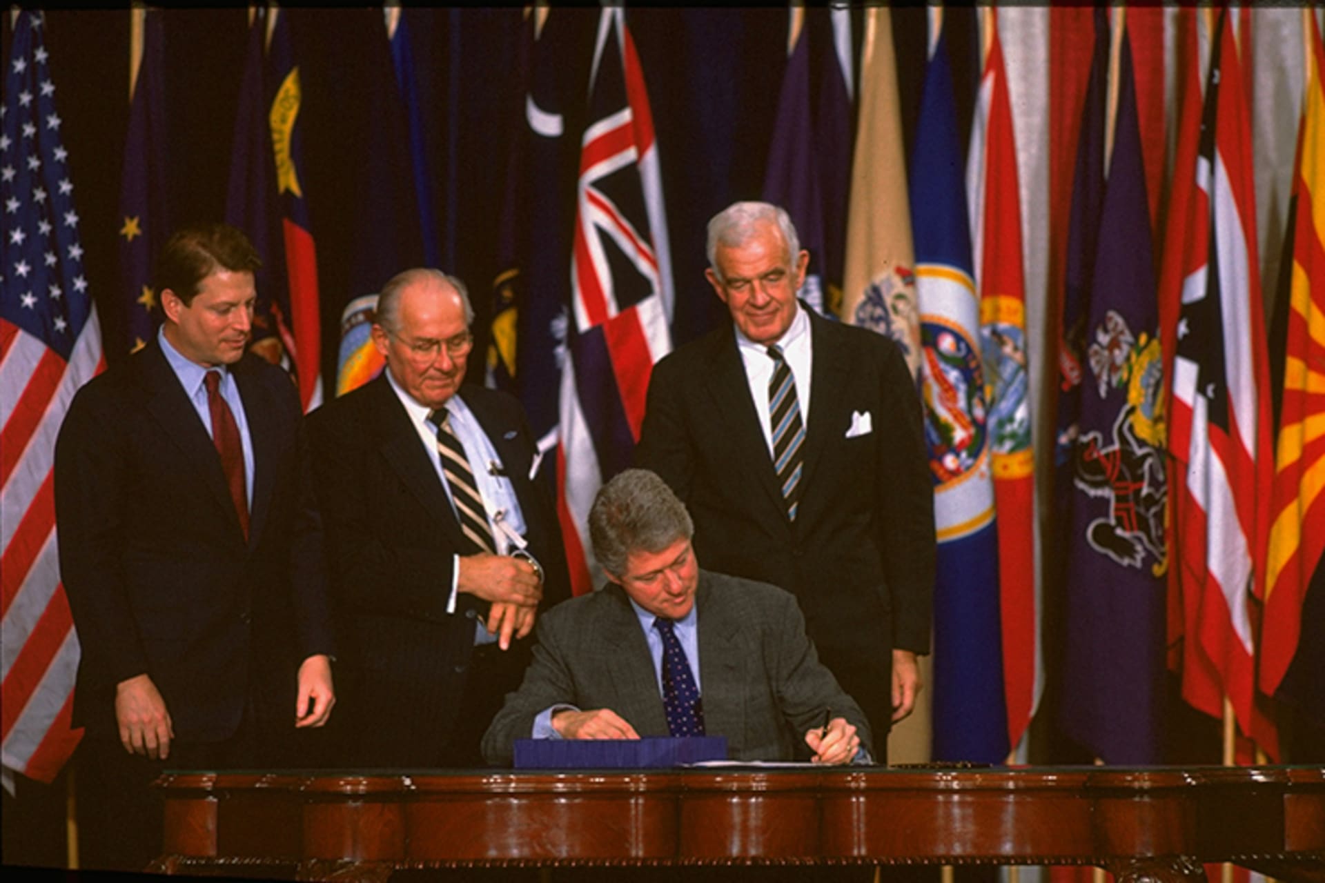 A photo of President Bill Clinton signing the NAFTA trade agreement as other members of government flank him. There are various countries flags in the background.