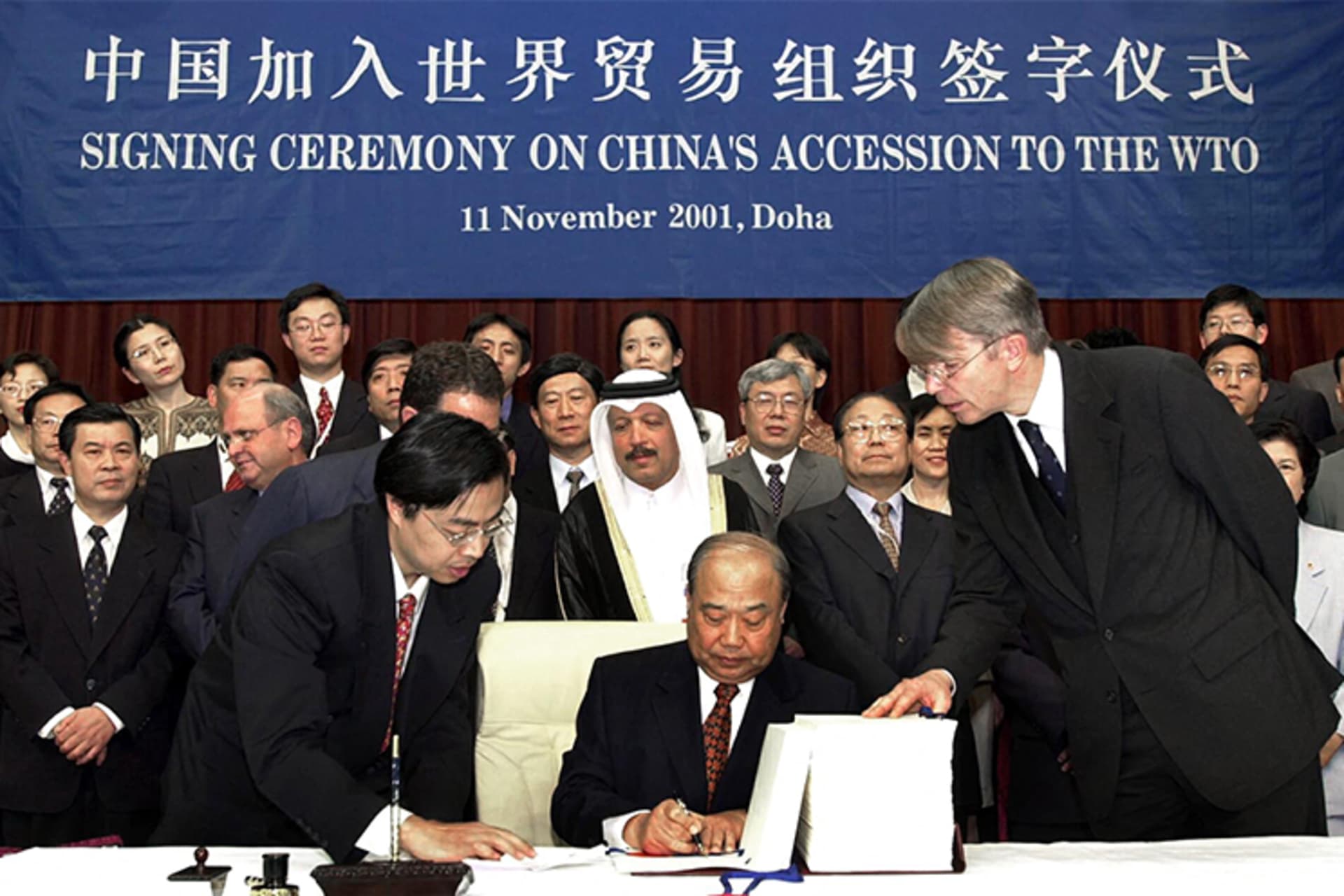A photo of Chinese Foreign Trade and Economic Cooperation Minister Shi Guangsheng signing China's WTO accession documents as people look on. There is a banner that reads "Signing Ceremony on China's Accession to the WTO" in the background in both Chinese and English. 