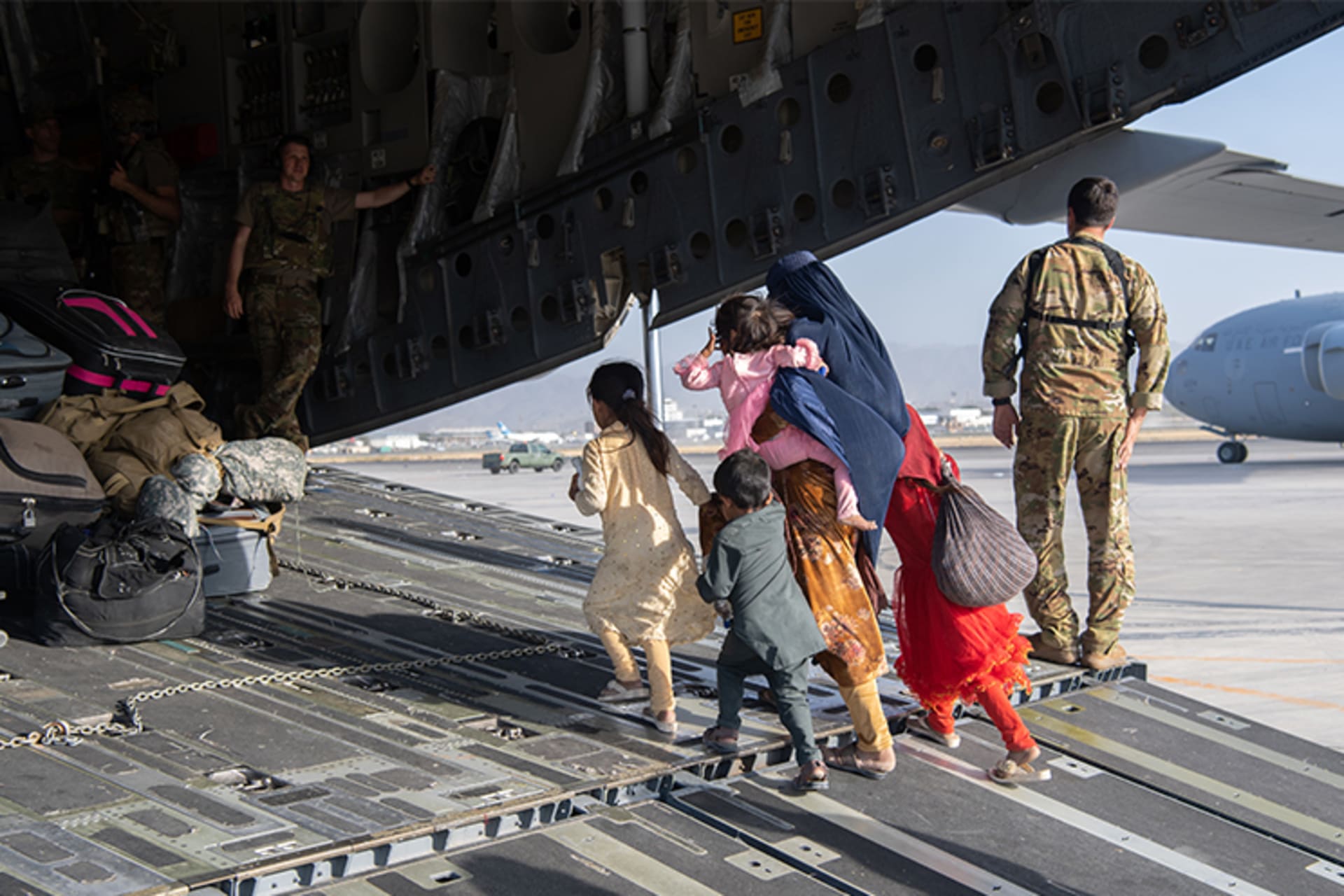 A photo of U.S. soldiers helping load passengers onto a U.S. plane at an airport as people evacuate. A woman with four kids walks on to the plan which has been loaded with many belongings. 