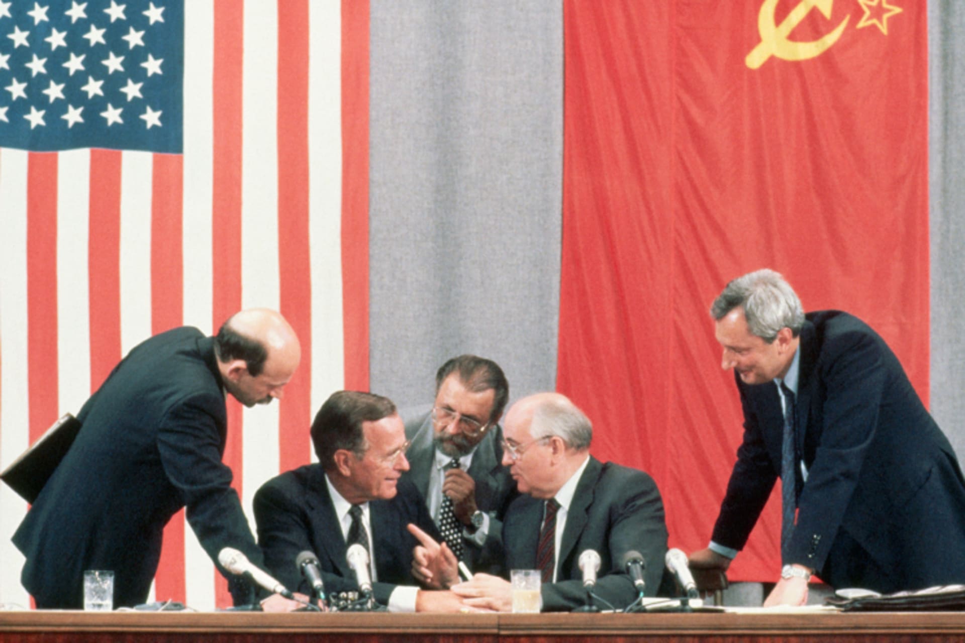 A photo of President George Bush and Soviet leader Mikhail Gorbachev during the 1991 Moscow Summit. The two converse as other men listen in. Behind them are the American and USSR flags hung on the wall.