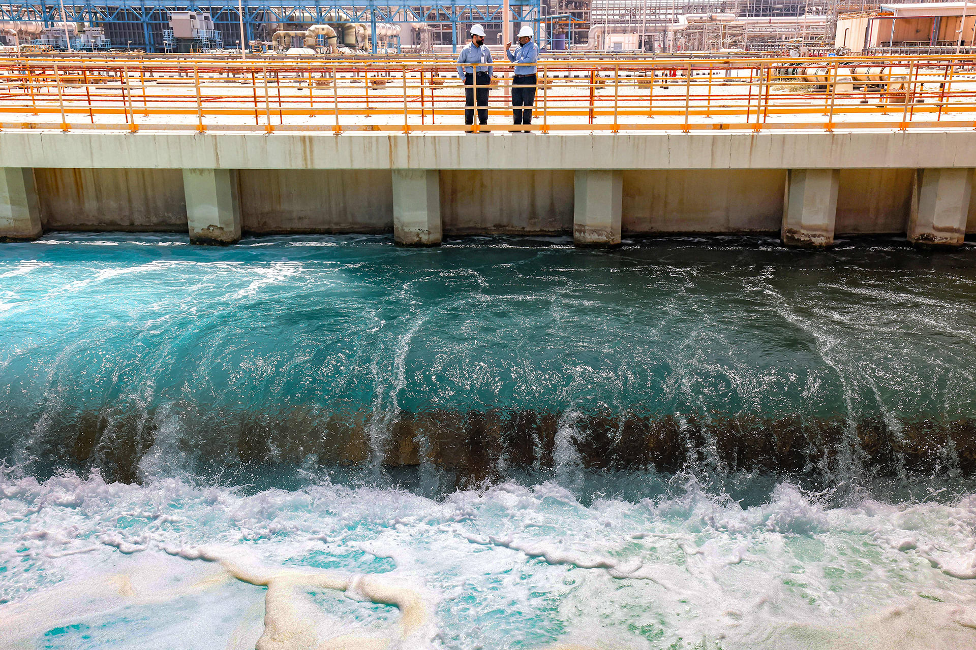 A manager at the Ras al-Khair water desalination plant speaks with an employee at the facility in Ras al-Khair along the Gulf coast in eastern Saudi Arabia. Water flows underneath them while they stand on a concrete platform.