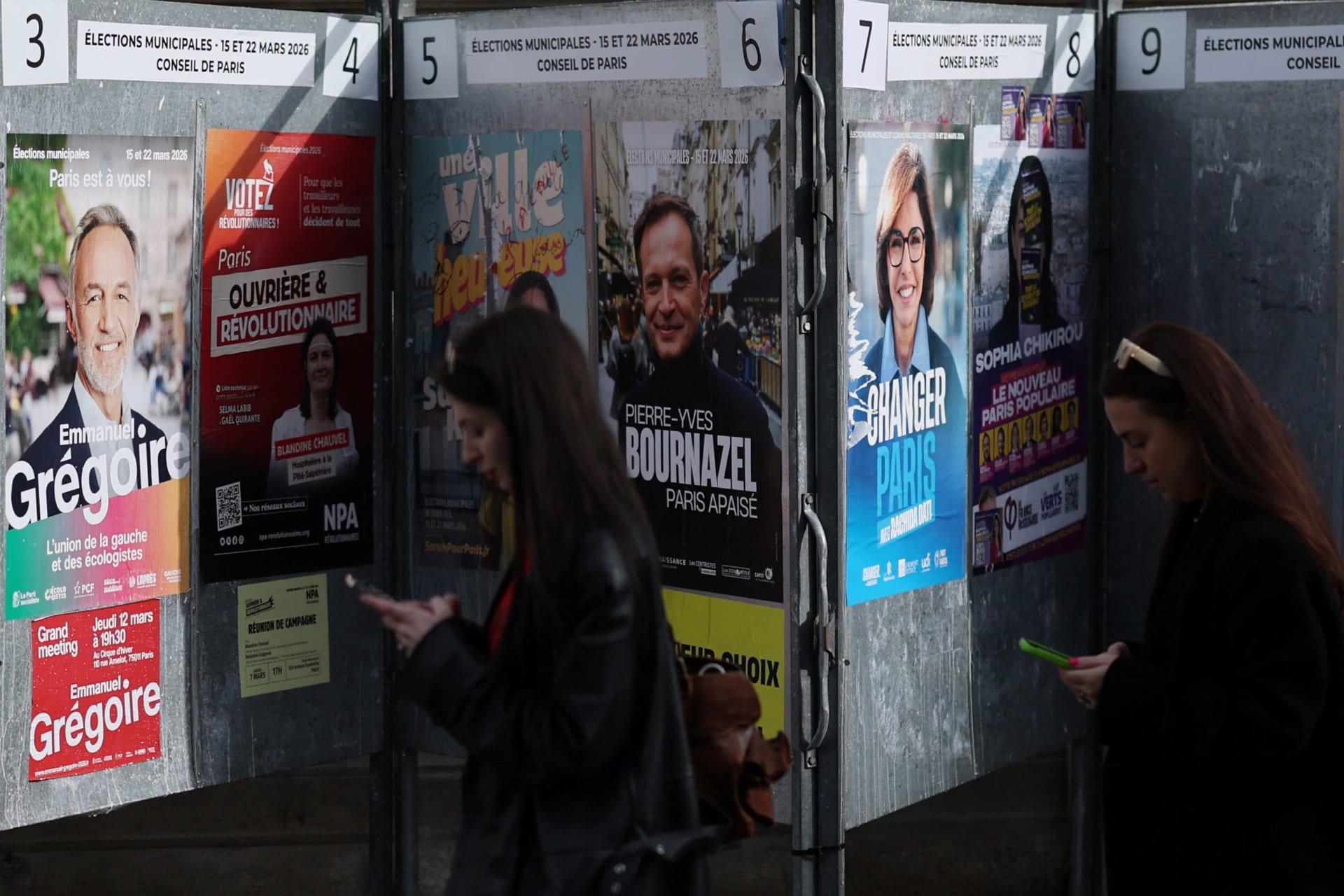 Women looking at their mobile phones walk past electoral campaign panel boards with posters of Paris mayoral election candidates ahead of the upcoming mayoral elections in Paris, France, March 6, 2026.