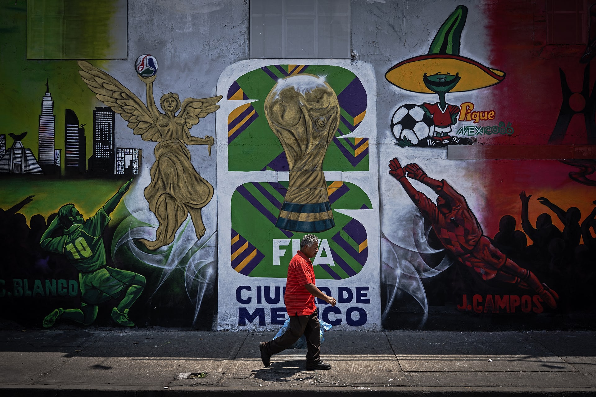 A man walks down a sidewalk in front of a concrete wall displaying a painted mural for the 2026 World Cup in Mexico.