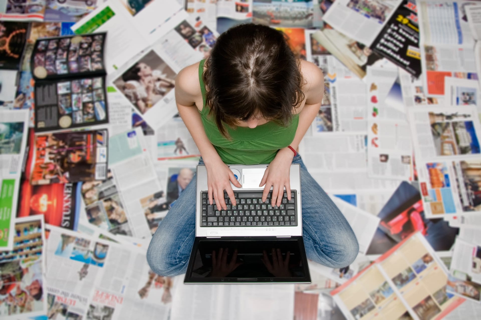 A woman sits on her computer typing on a floor covered with newsletters