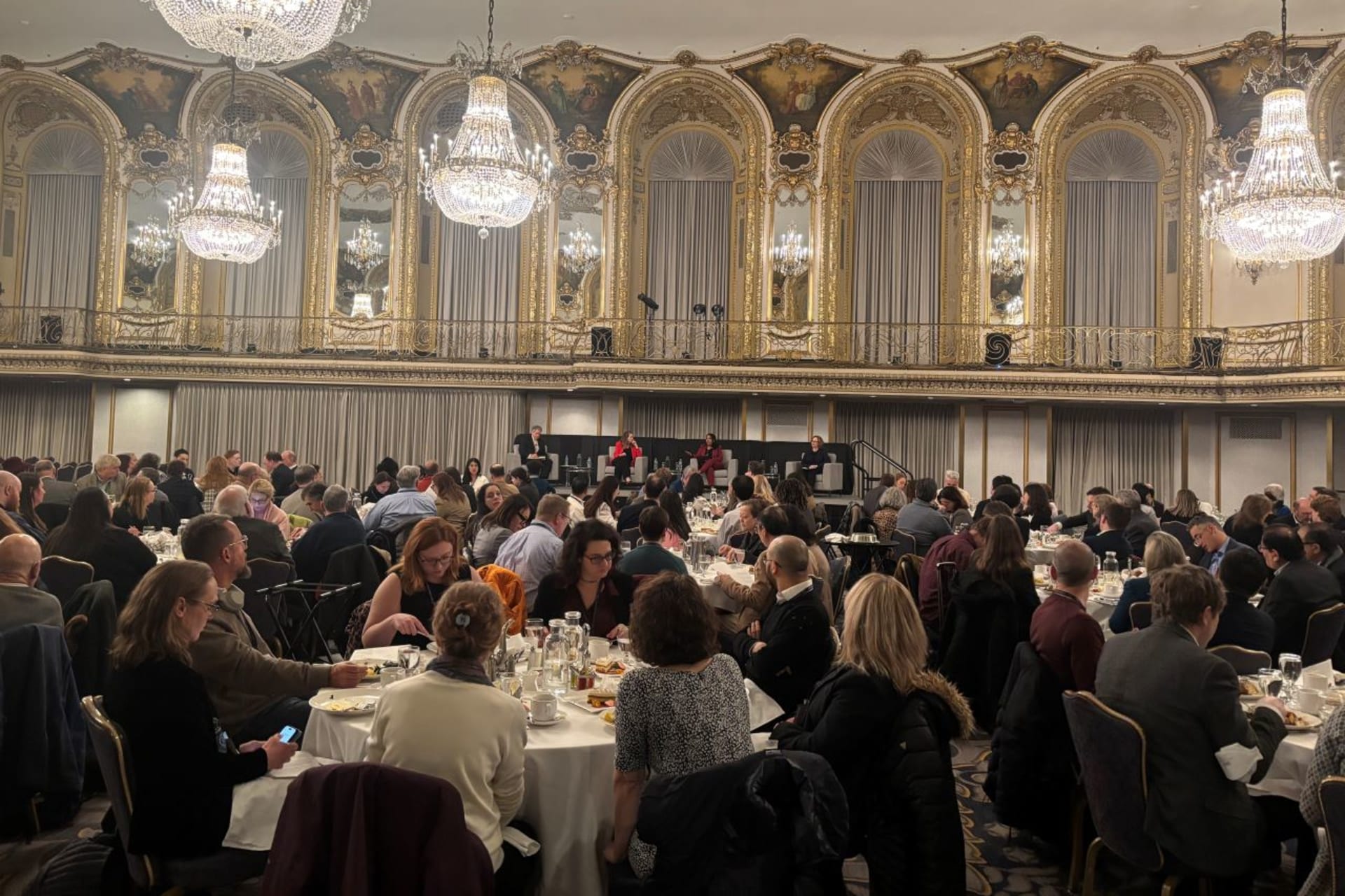 A group of professors sit in rounds eating lunch, while four speakers are on a lifted stage.