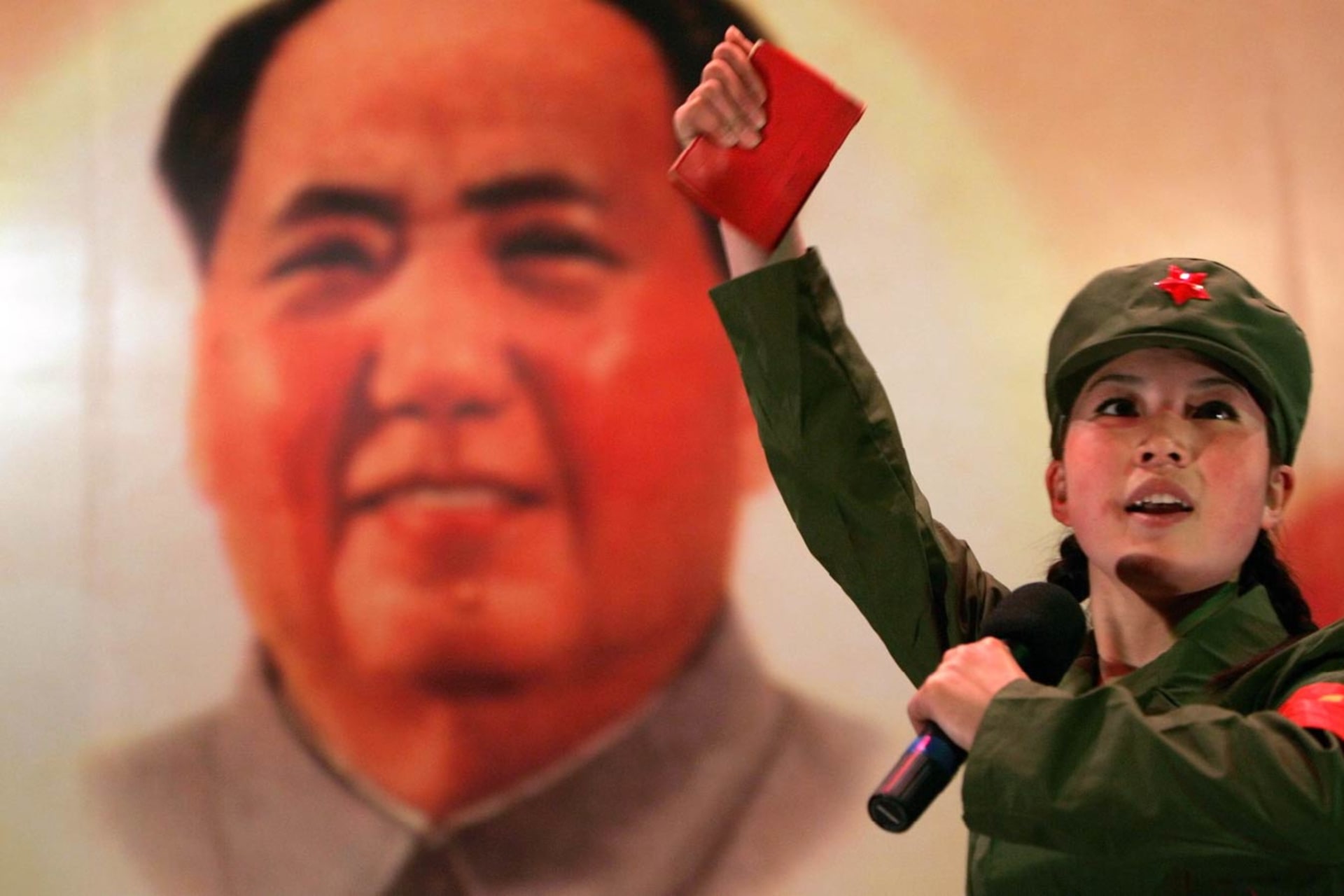 A Chinese woman dressed in a Red Guard uniform holds a little red book in her extended hand and a mic in the other. A painting of Mao is behind her.