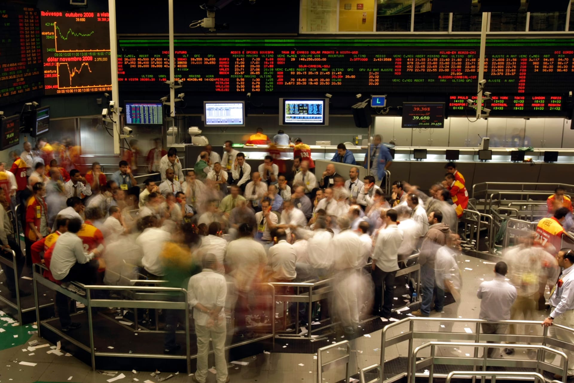 Traders are gathered, working on the main floor of a stock exchange market. There are boards showing stock values behind them.
