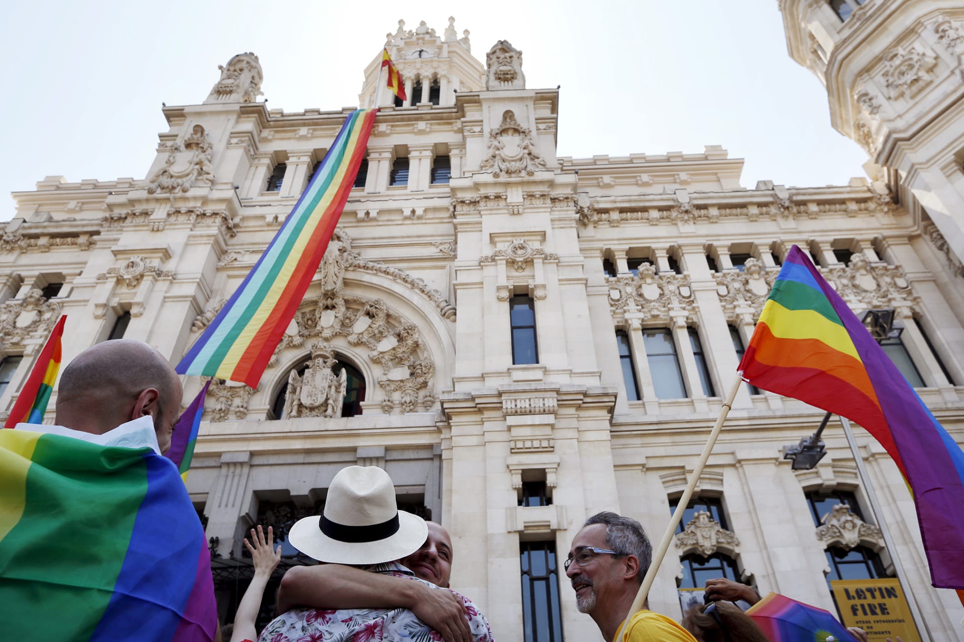 People celebrate pride with flags, rainbows, and hugs as a long rainbow flag is displayed from the Town Hall in Madrid.