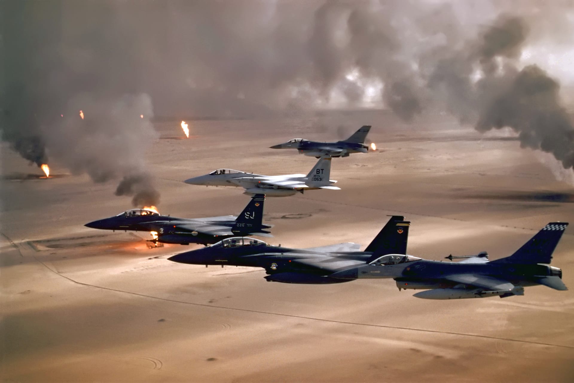 Photo showing U.S. fighter jets flying over a desert landscape spotted with oil well fires spewing plumes of smoke into the air.