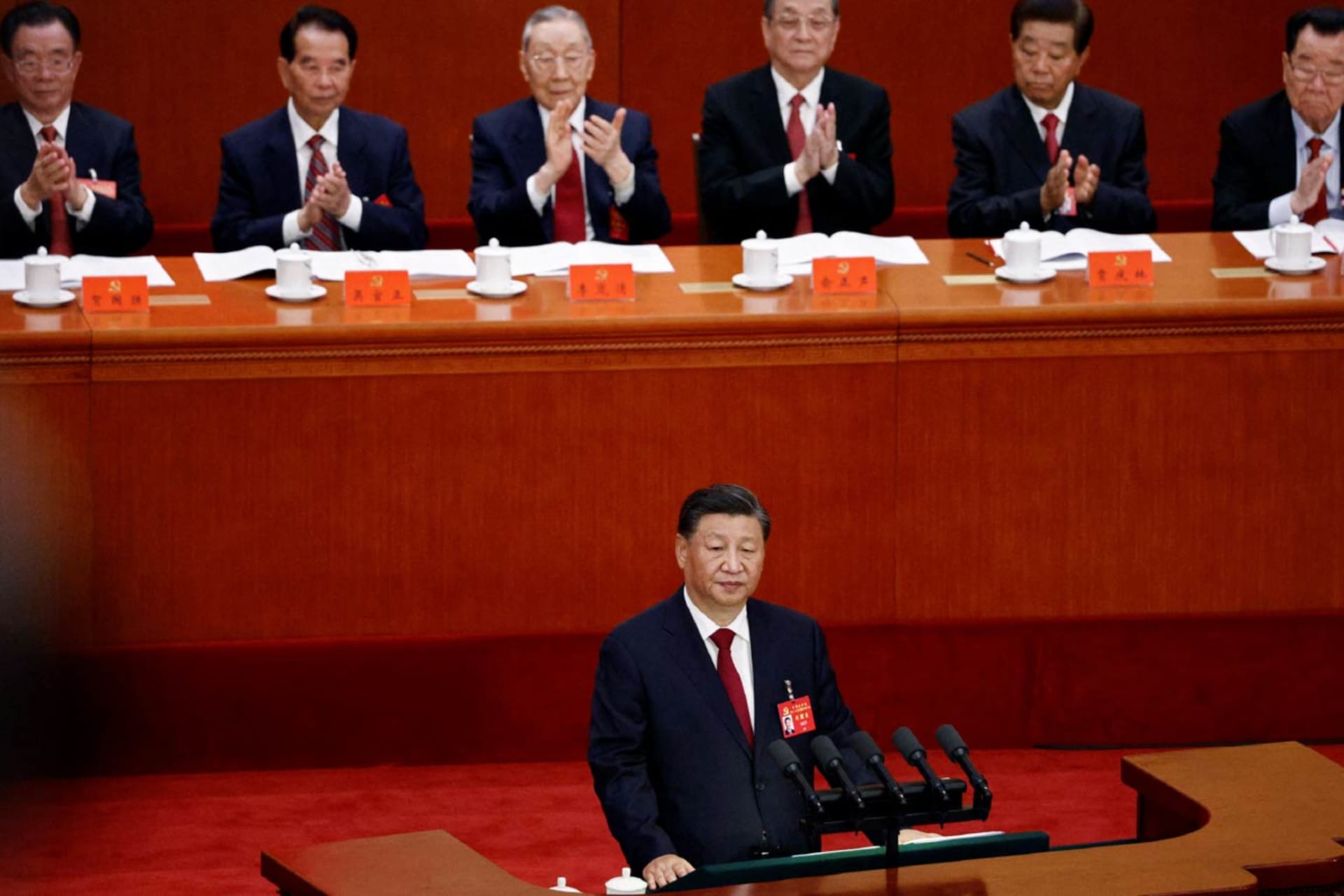 Chinese President Xi Jinping stands at a podium to give a speech. Other Chinese men sit behind him and applaud.