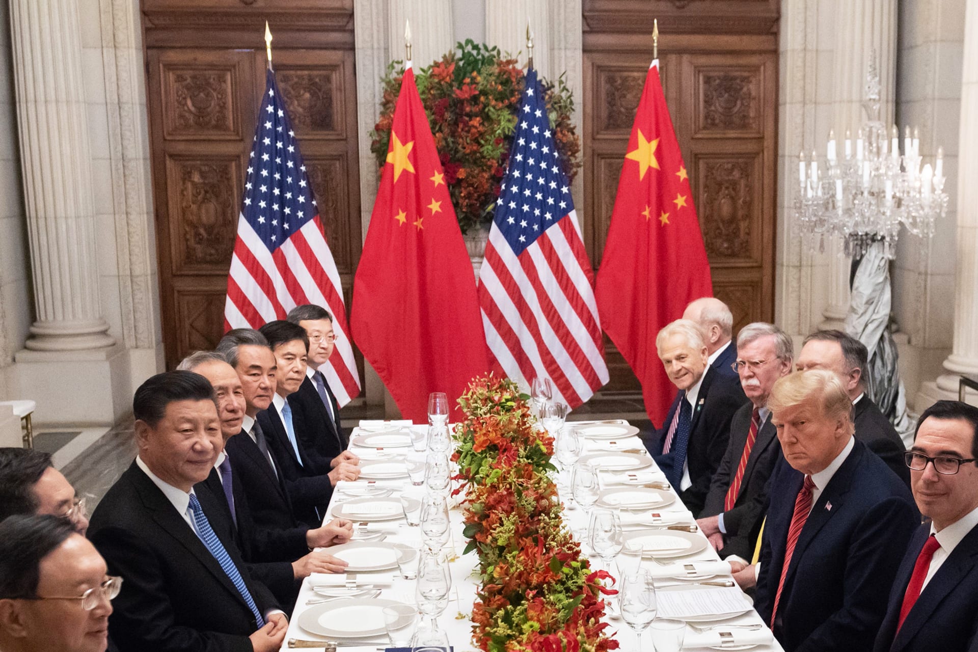 U.S. President Donald Trump and China's President Xi Jinping hold a dinner meeting. The two presidents and other men are seated on opposite sides of a long table as the American and Chinese flags can be seen in the background.