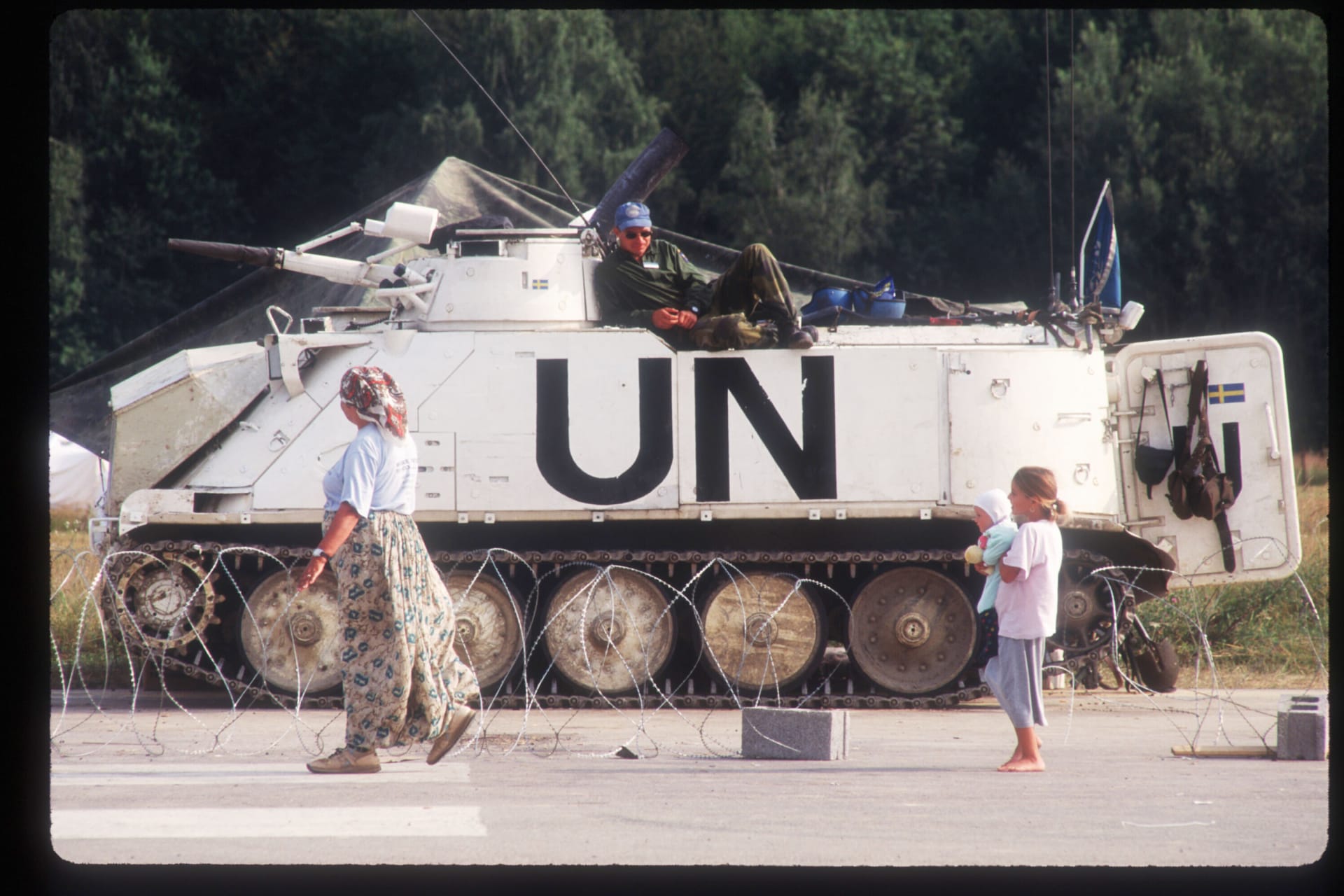 Refugees, a woman and young girl holding a baby, walk near a UN tank as a man watches them from atop the tank.