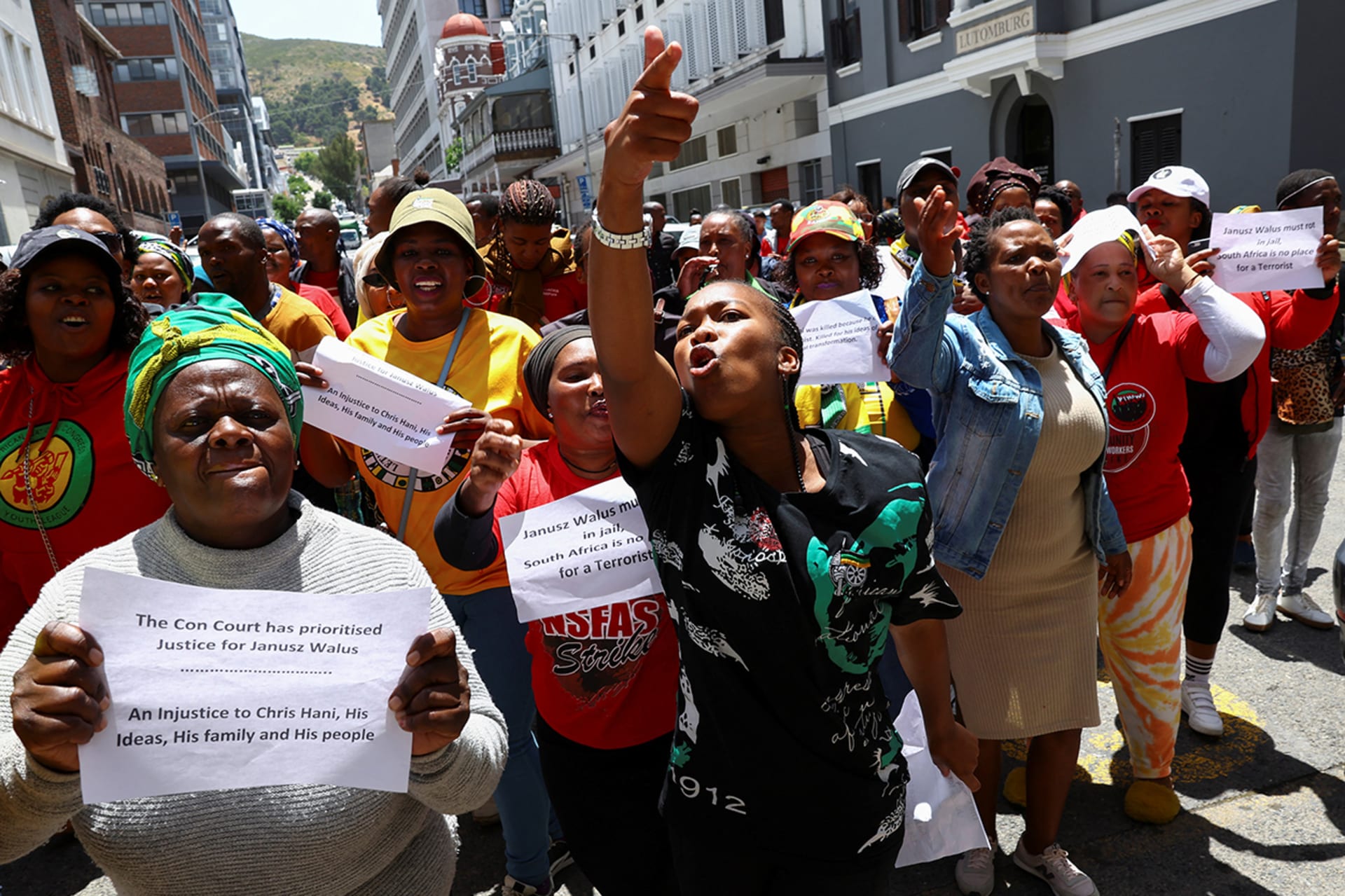 People demonstrate outside. One demonstrator raises their hand and points a finger, while another holds a paper that reads "The Con Court has prioritised Justice for Janusz Walus. An Injustice to Chris Hani, His Ideas, His family and His people".
