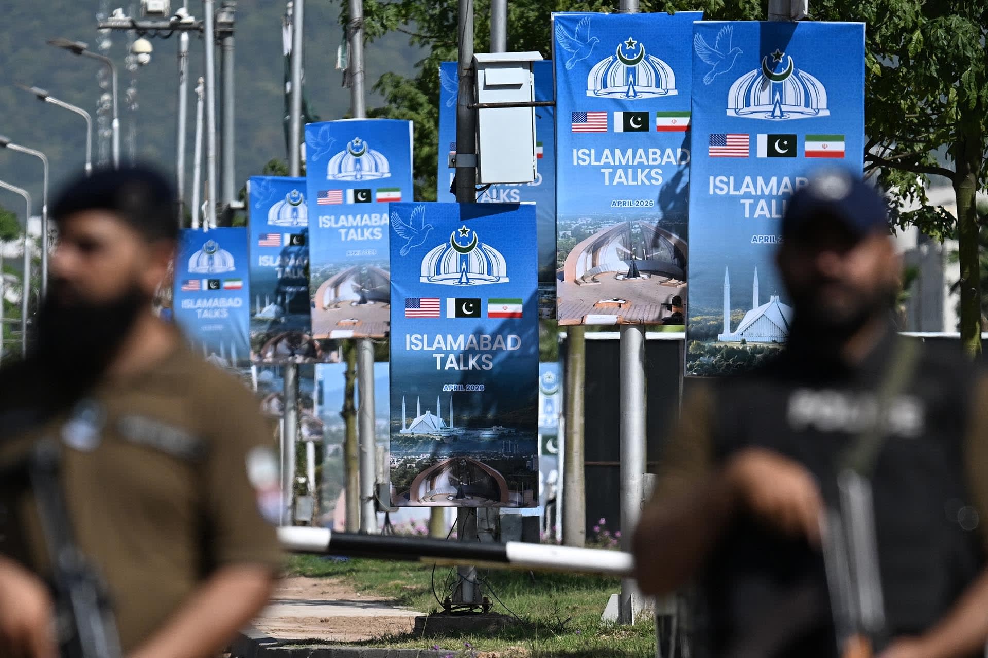 Two security guards stand armed in front of a road that has several banners displaying signage that read "Islamabad talks April 2026".