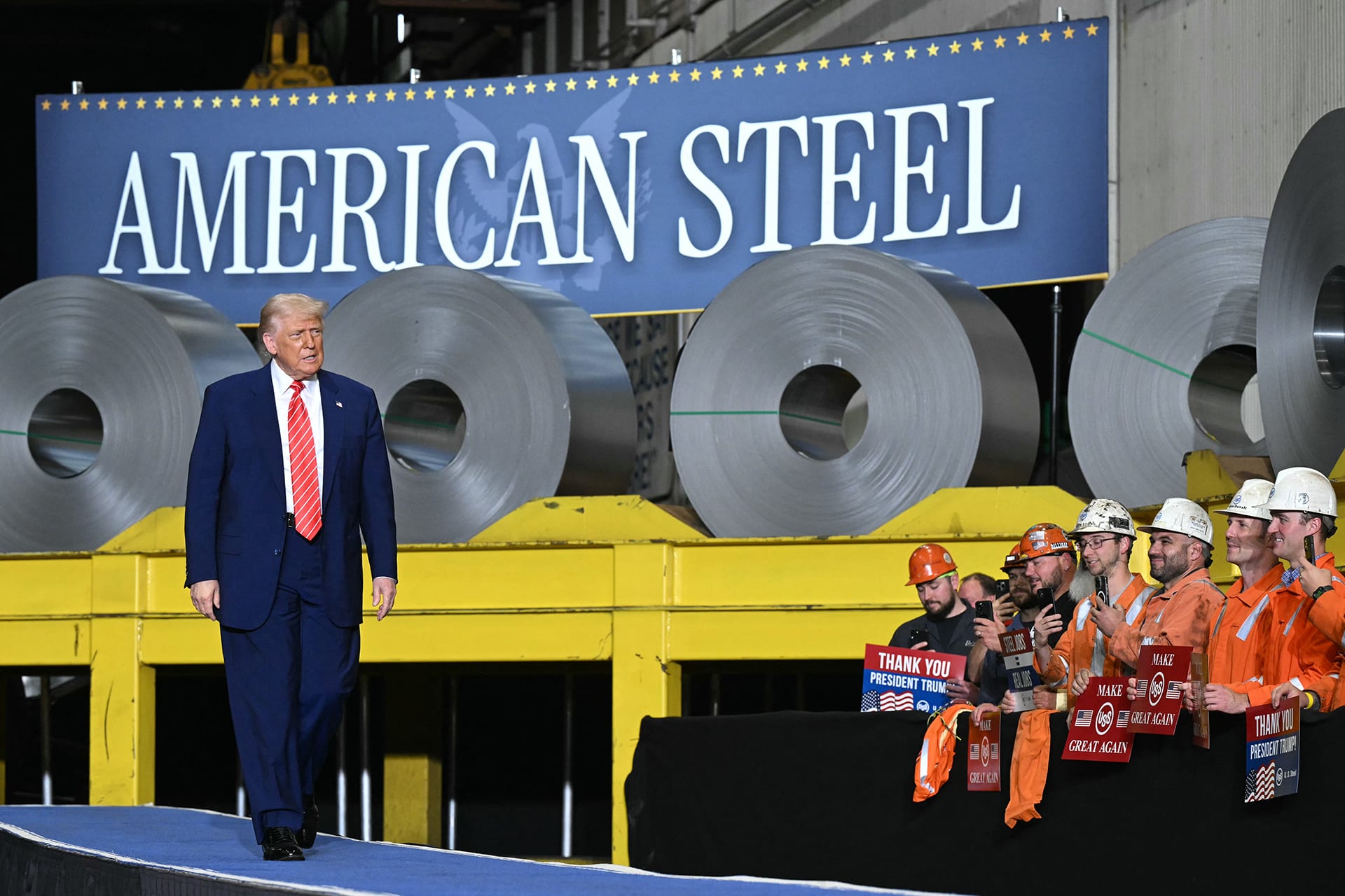 Photo of president trump standing in front of a banner and a pile of steel rolls to mark deal between Nippon Steel and US Steel