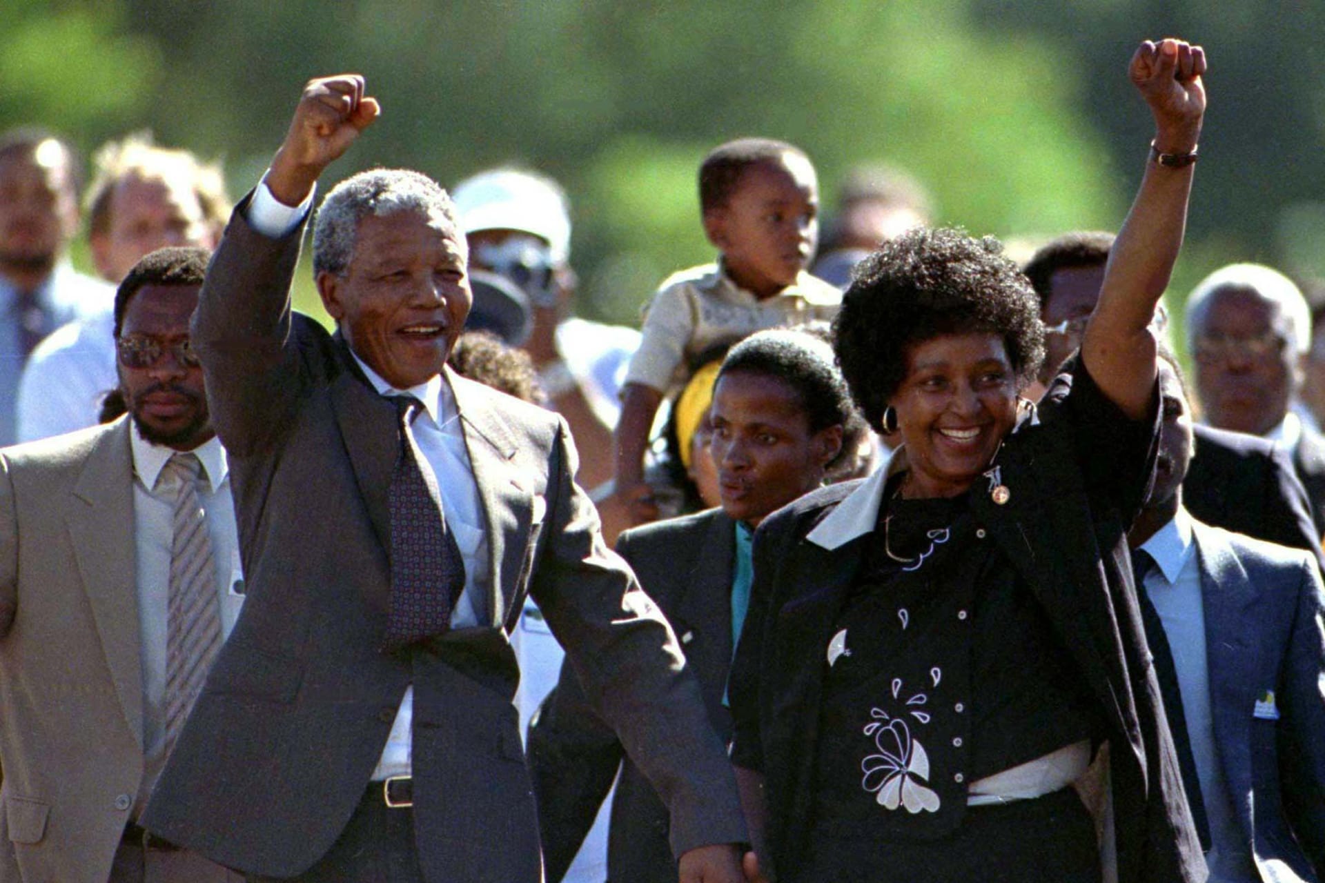 Nelson Mandela, accompanied by his wife Winnie, walks out of of prison. Both of them raise their fists in the air triumphantly as people walk behind them.