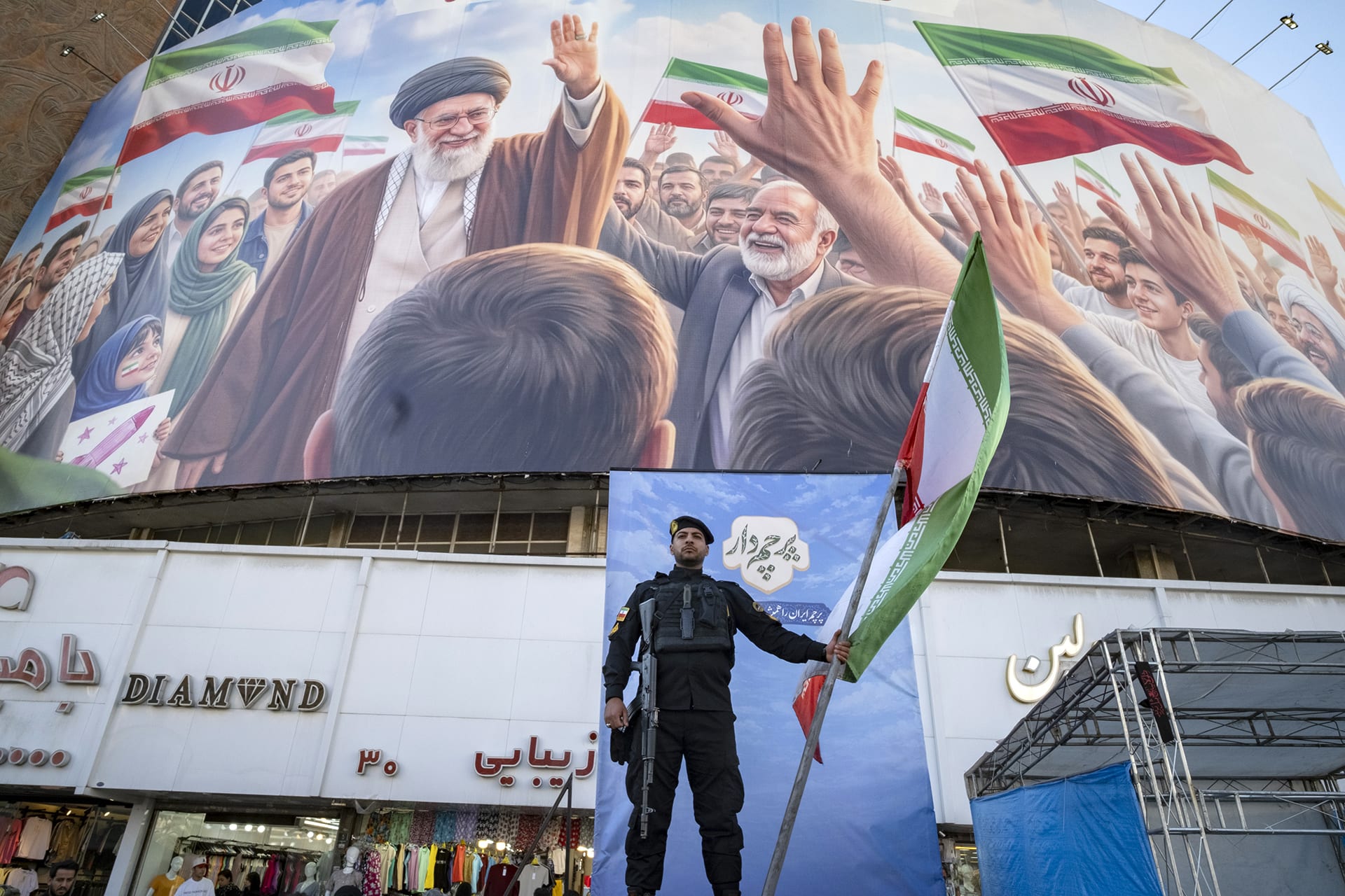 An Iranian police officer holds a national flag on a stage alongside a Persian script reading 'Flag bearer', under a giant billboard depicting an AI-generated portrait of Iran's late Supreme Leader Ayatollah Ali Khamenei in downtown Tehran, Iran, on April 23, 2026.
