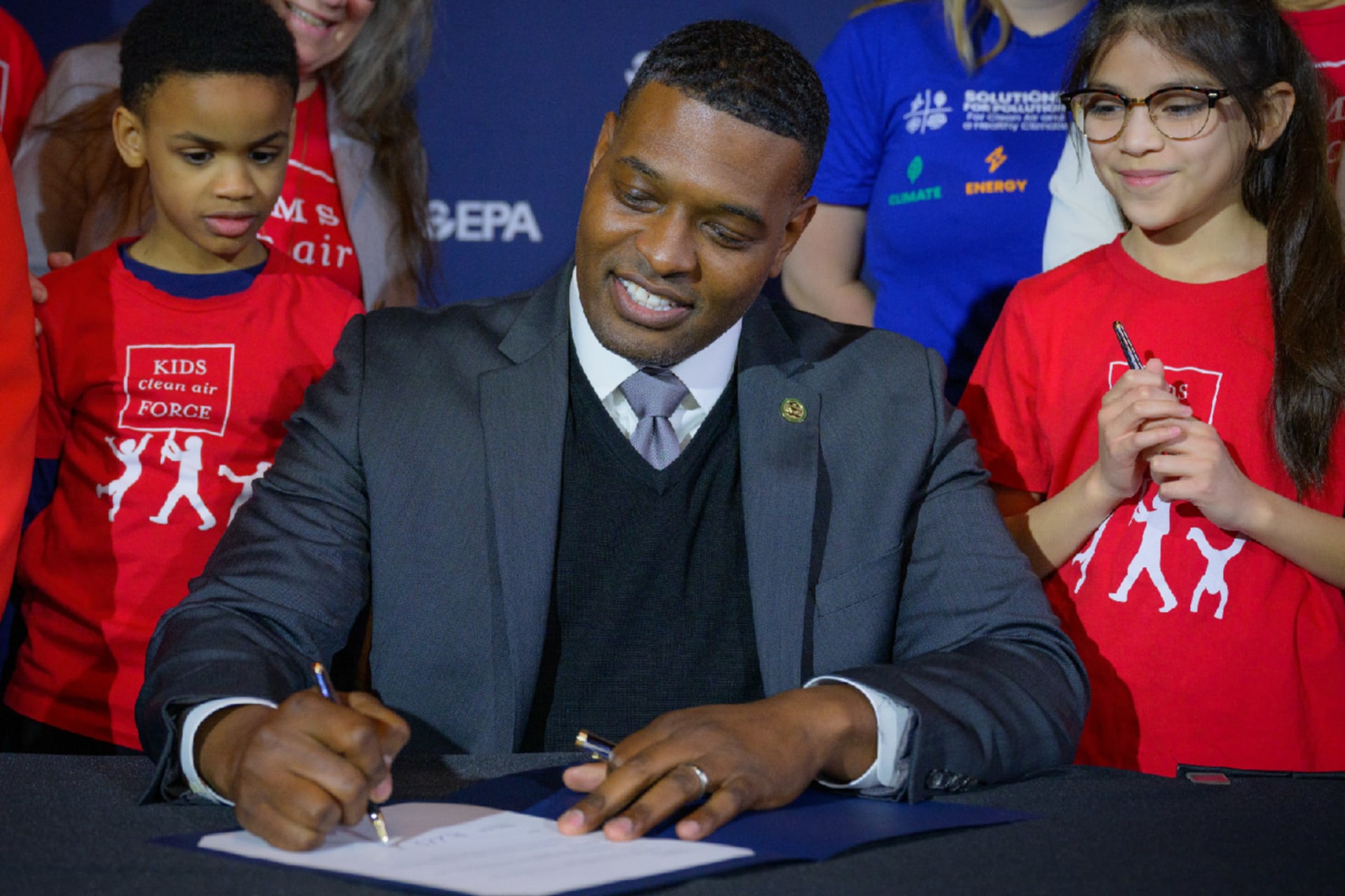 Environmental Protection Agency Administrator Michael S. Regan signs a paper during a ceremonial signing for stronger air quality standards. Two kids stand behind him wearing shirts that say "KIDS clean air FORCE".
