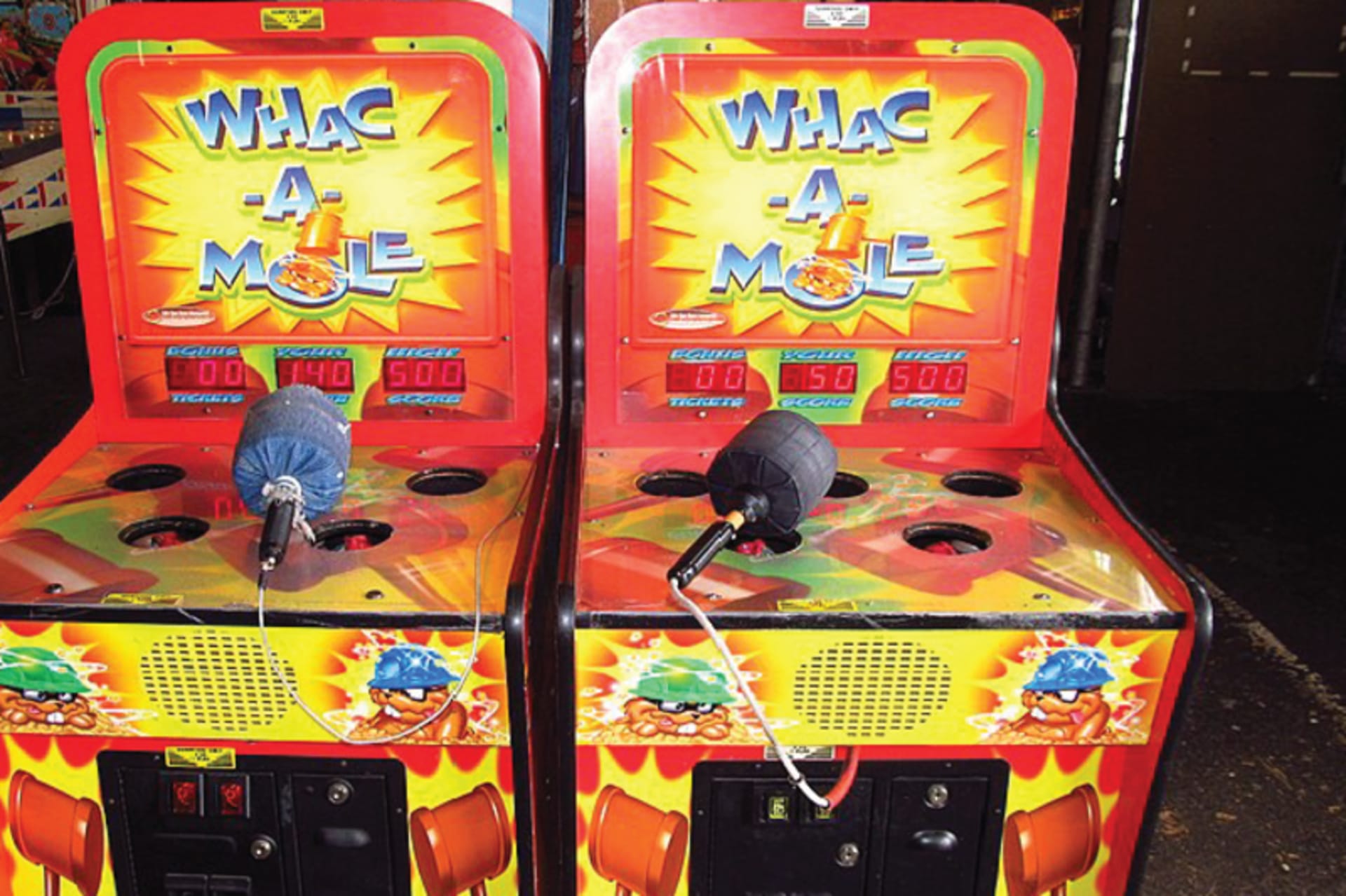 A picture of two Whac-A-Mole games at an arcade. In this game a player uses a soft hammer to hit plastic moles that randomly pop up from holes in a cabinet. Each time a mole is knocked down, another appears in a new hole.