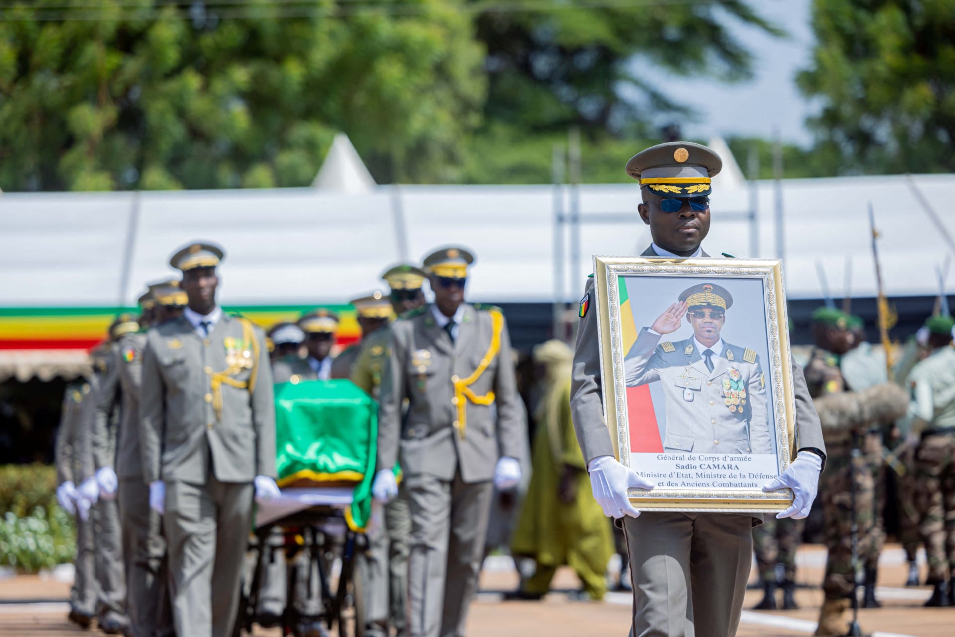 Men in military uniforms march carrying a coffin while another man in a uniform holds a framed photo.