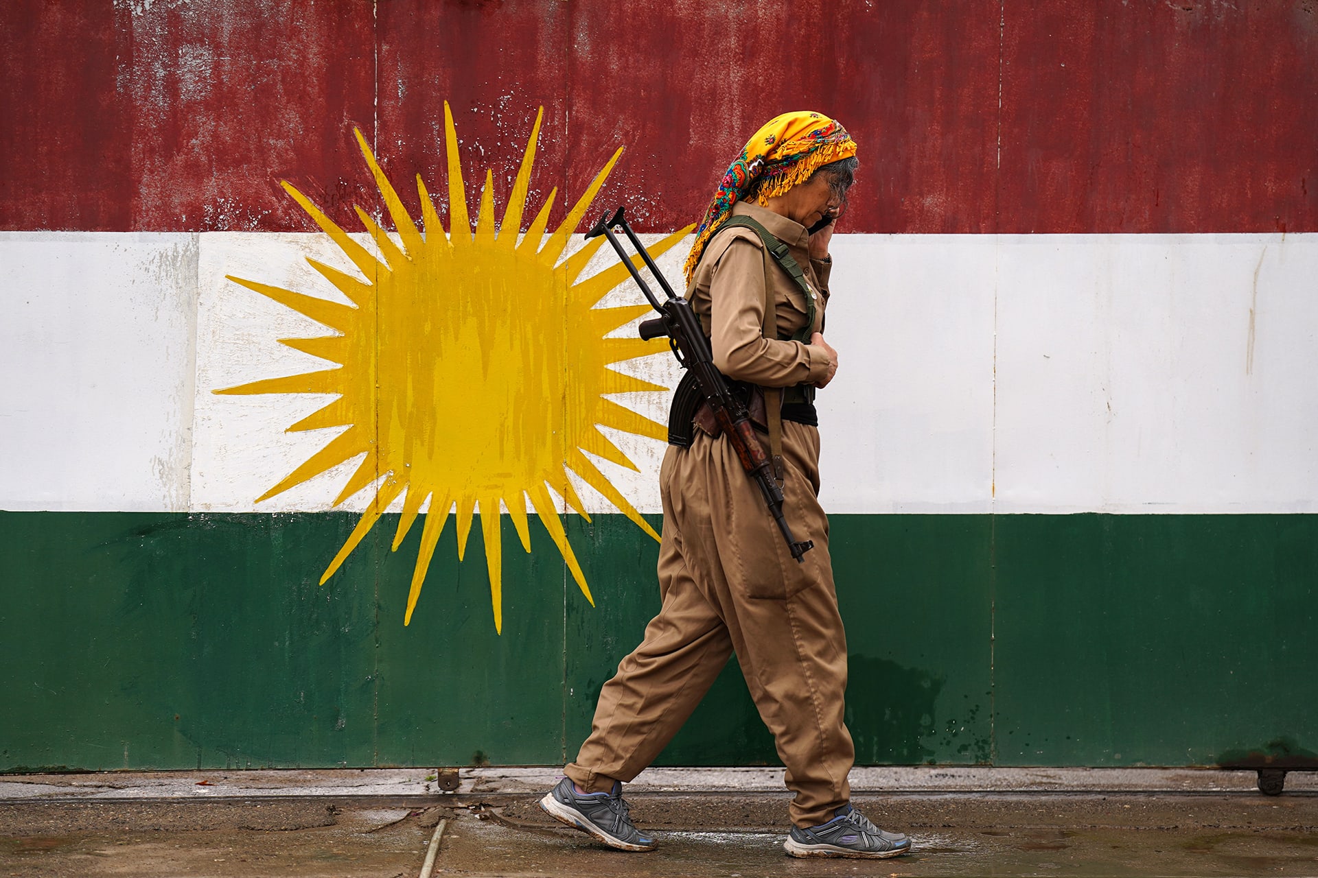 A female soldier walks in front of a Kurdish flag while on the phone