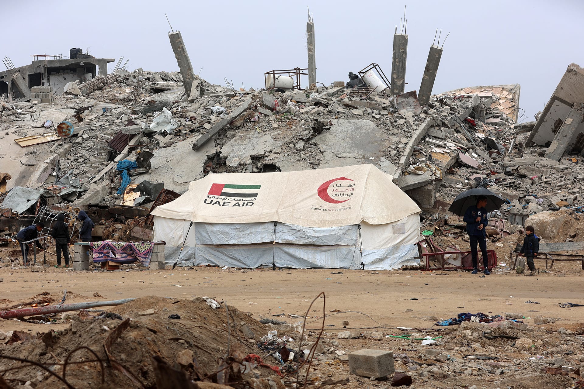 An aid tent stands on the dirt in front of a large pile of rubble and debris.