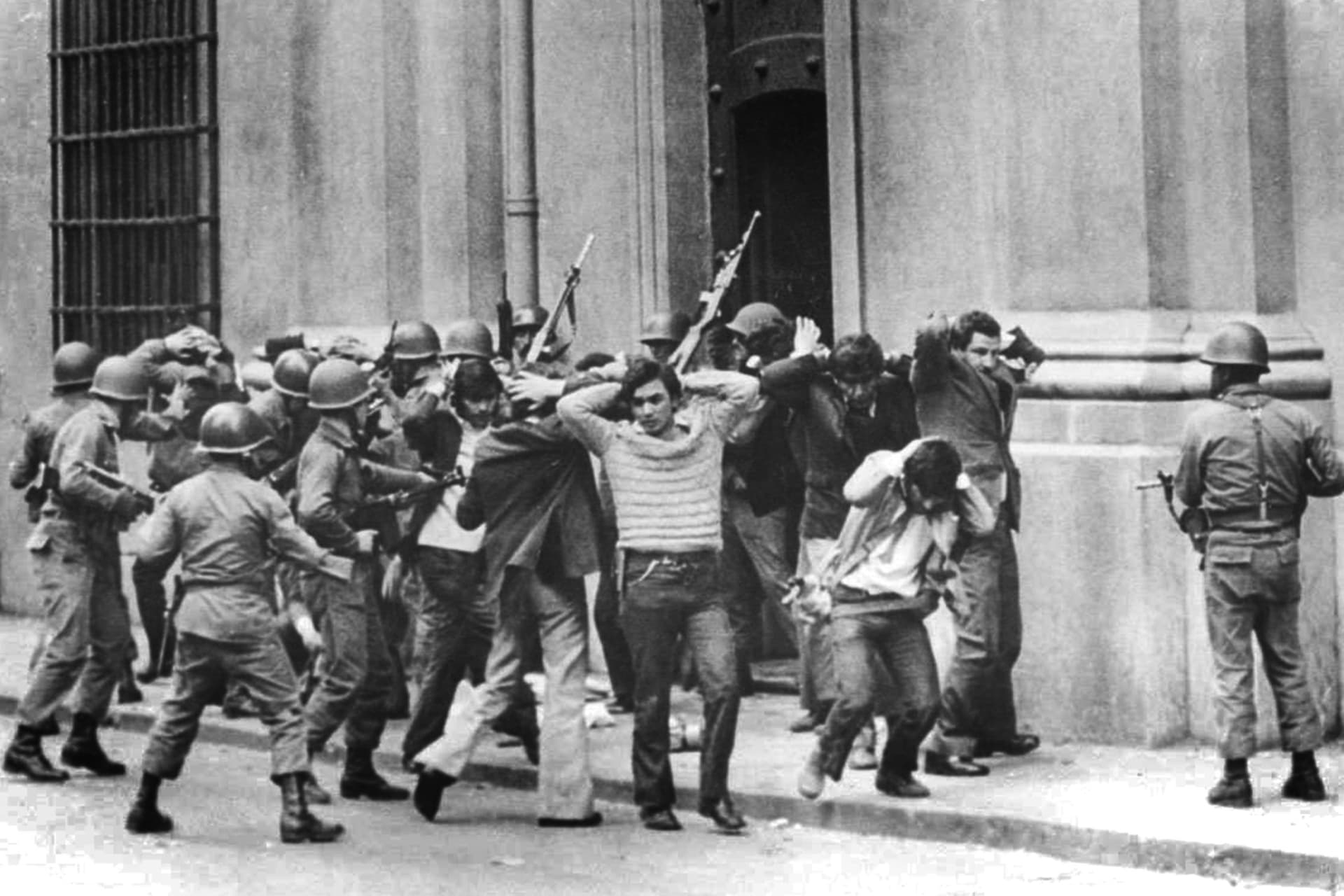 People with their hands up being led out by soldiers holding guns.