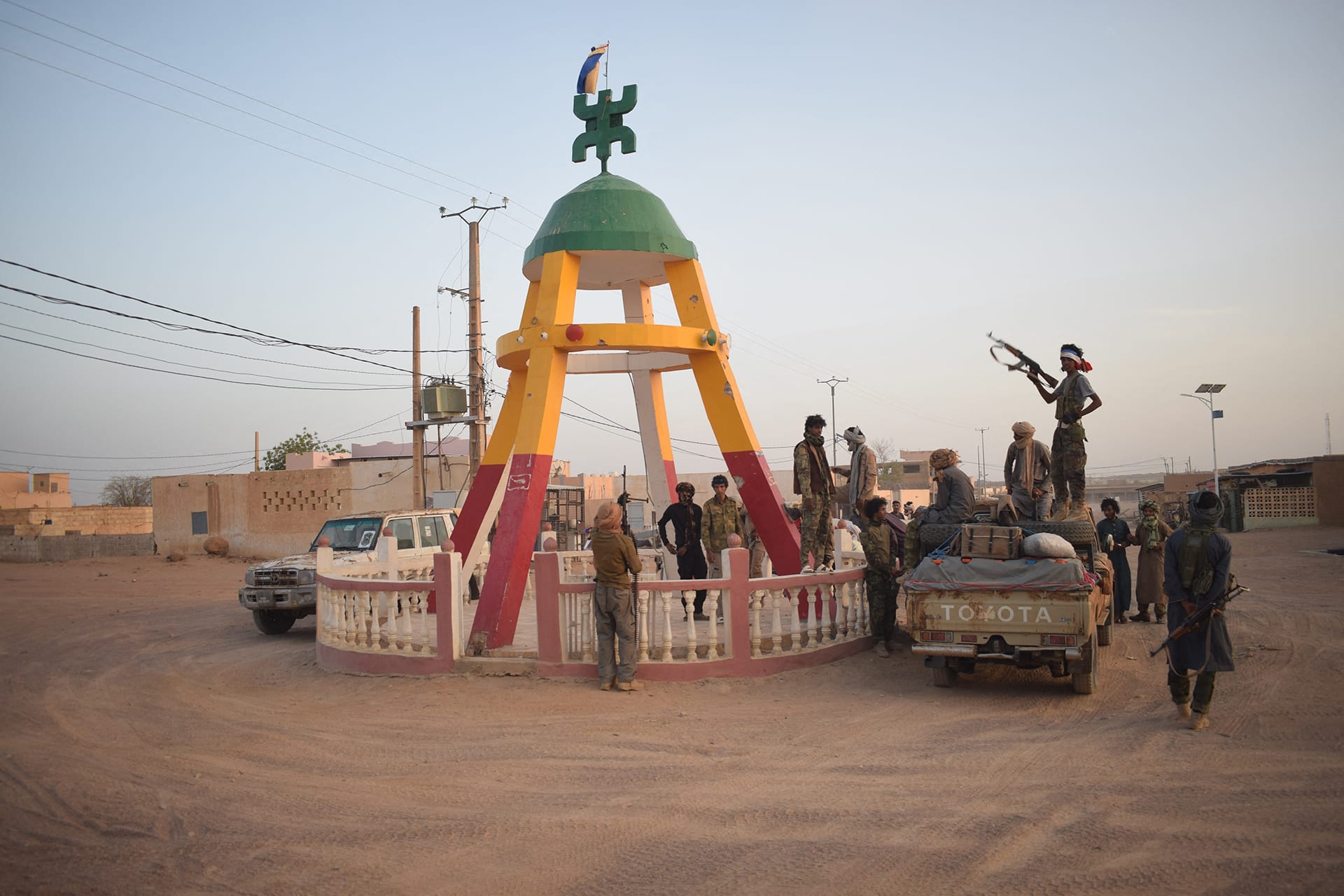 Soldiers gather on the dirt in front of a red, yellow, and green monument, with one soldier standing with his gun pointed up while on top of a truck.