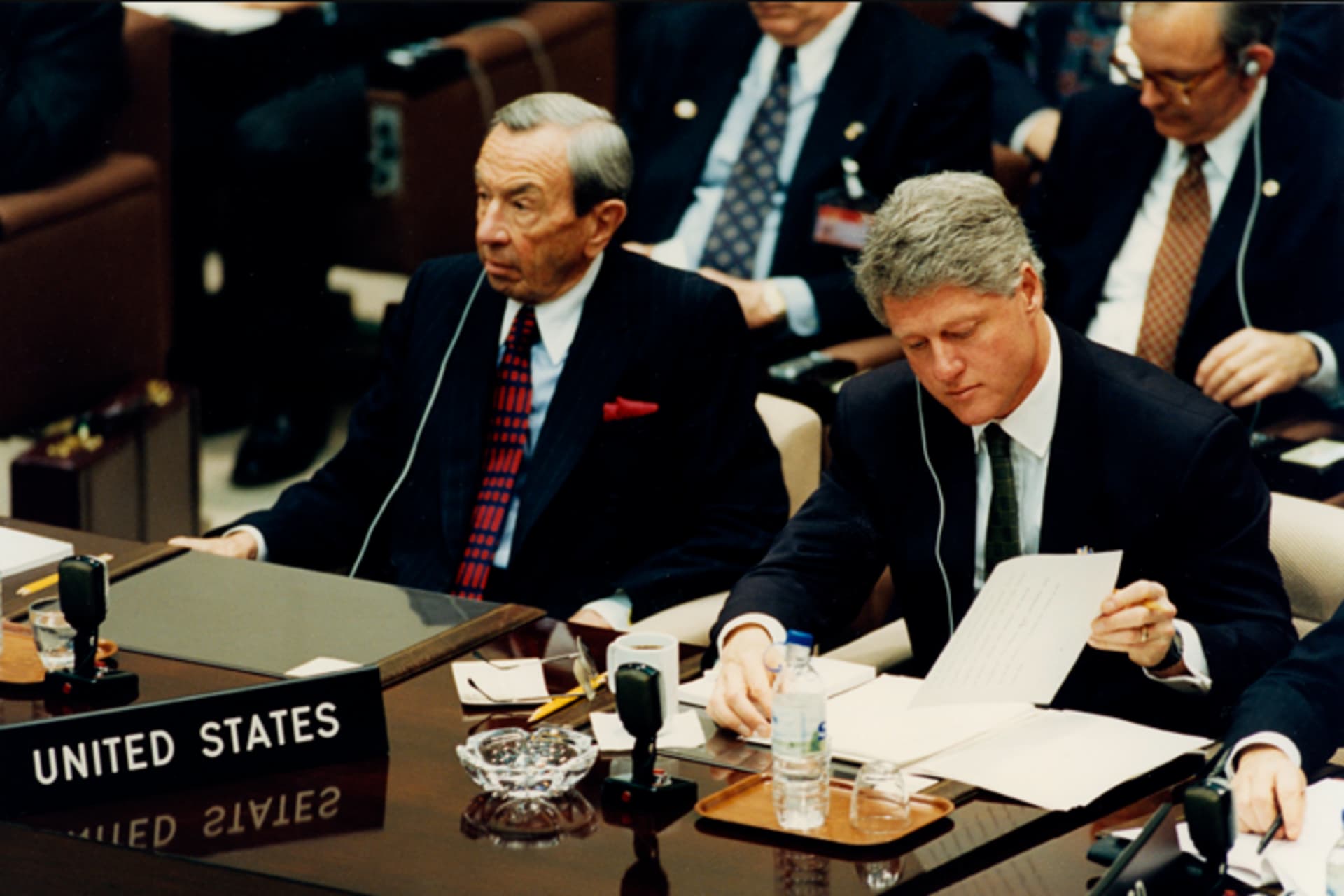 President Bill Clinton and Secretary of State Warren Christopher are seated next to one another. Clinton writes something on a piece of paper.