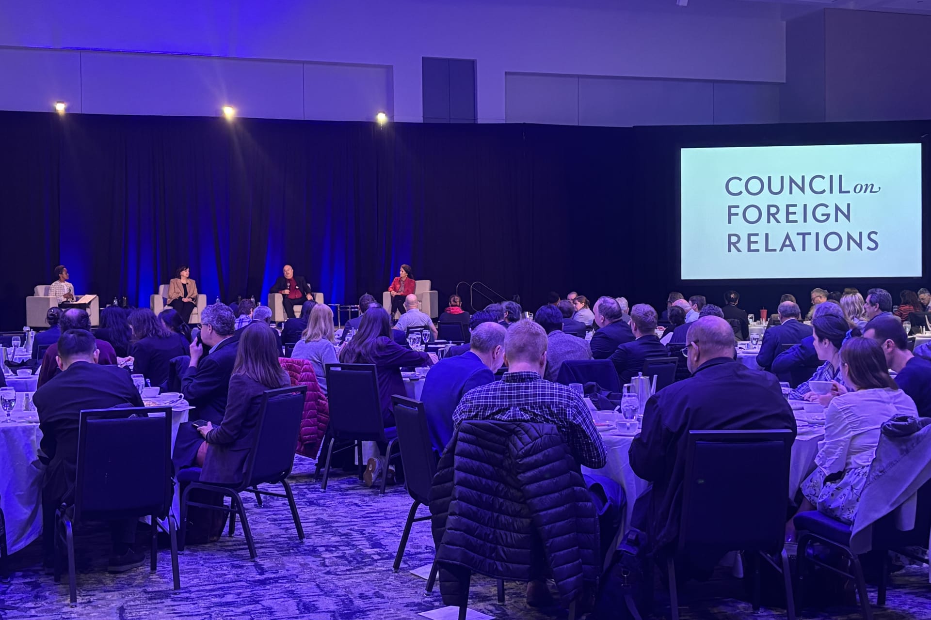 Four speakers on a raised stage with people in the audience eating lunch at rounds.
