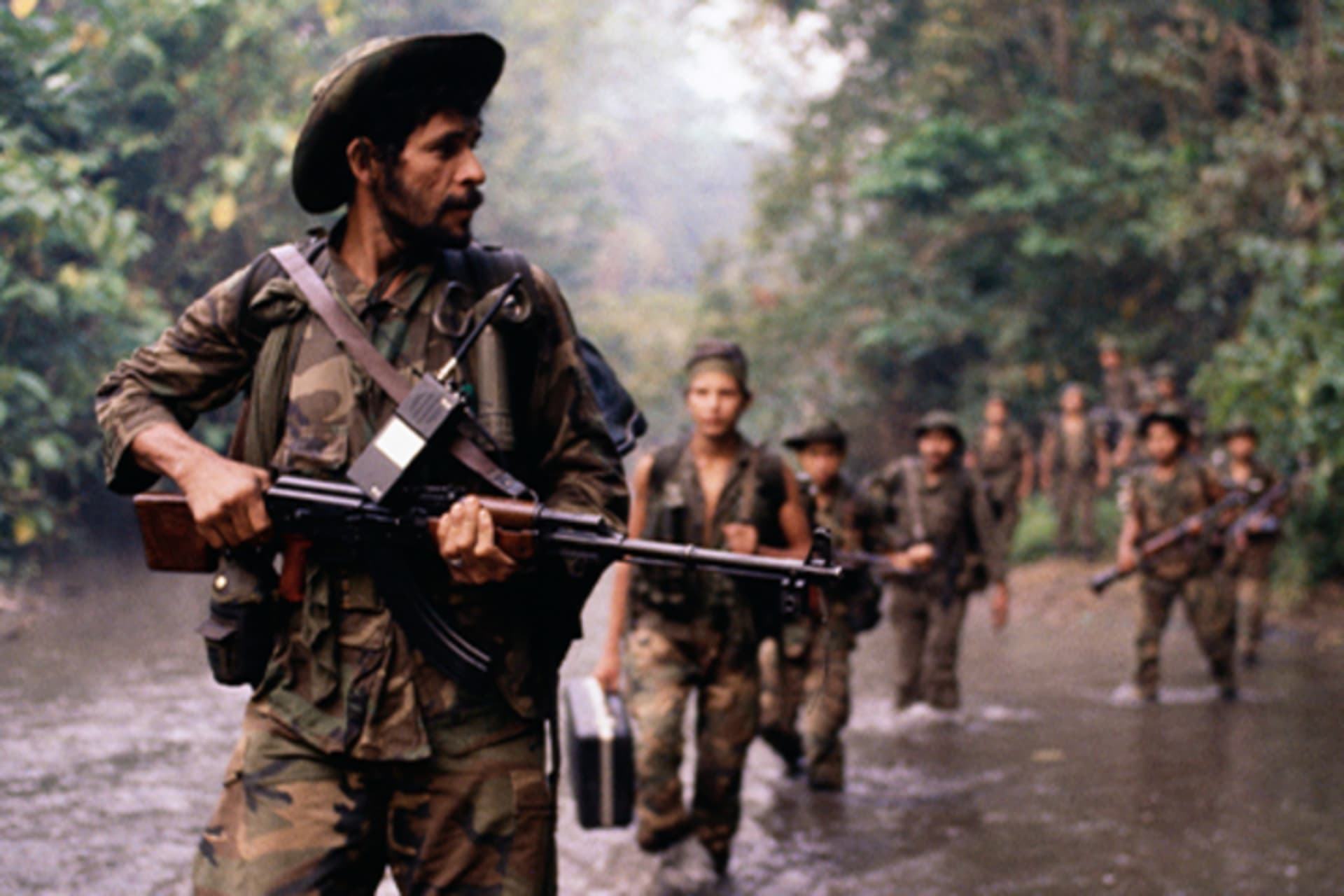 Men in military fatigues march with rifles in a waterway in a jungle.